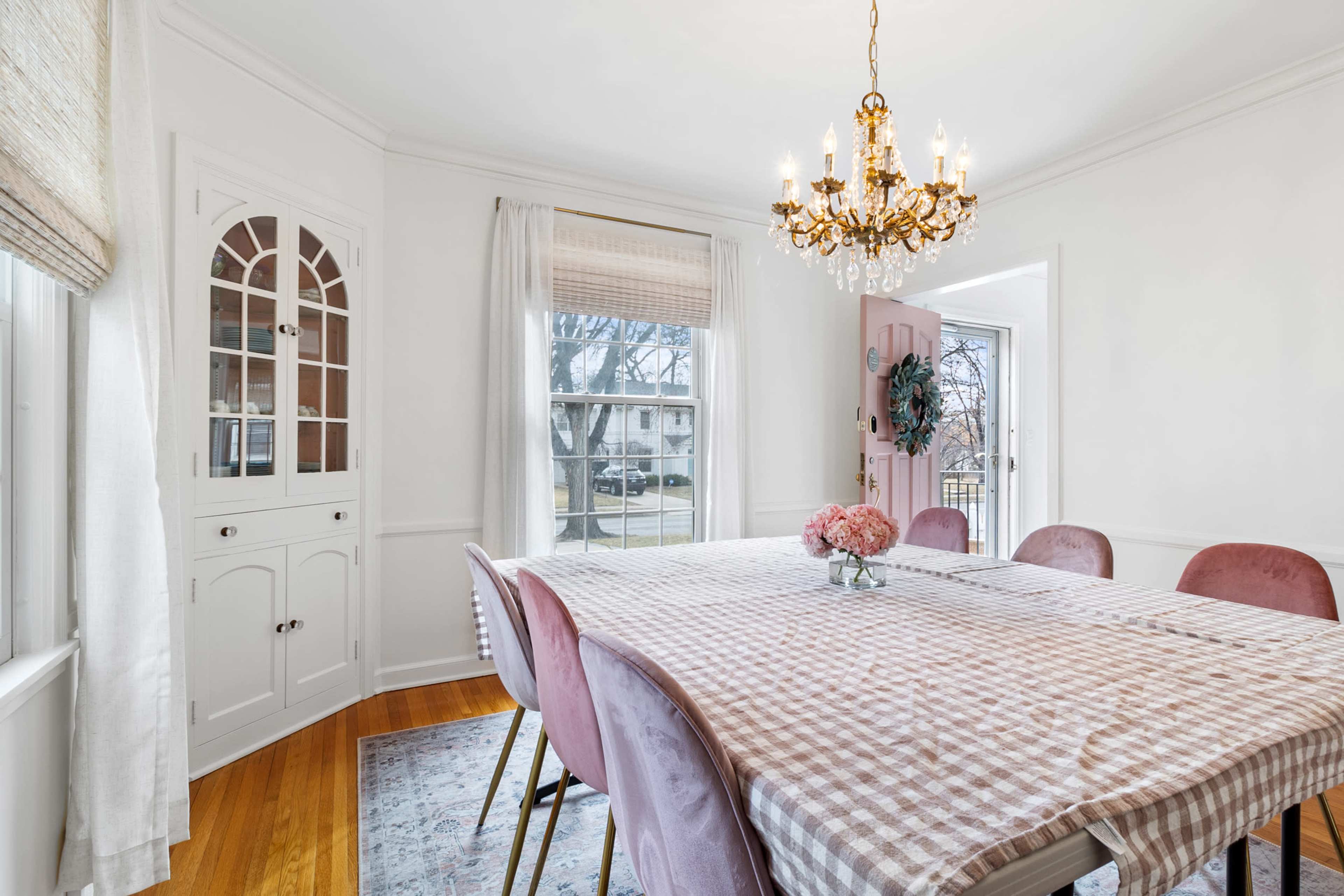 A dining room features a large table covered with a checkered tablecloth, surrounded by pink chairs and illuminated by a chandelier, with a cabinet and windows in the background.