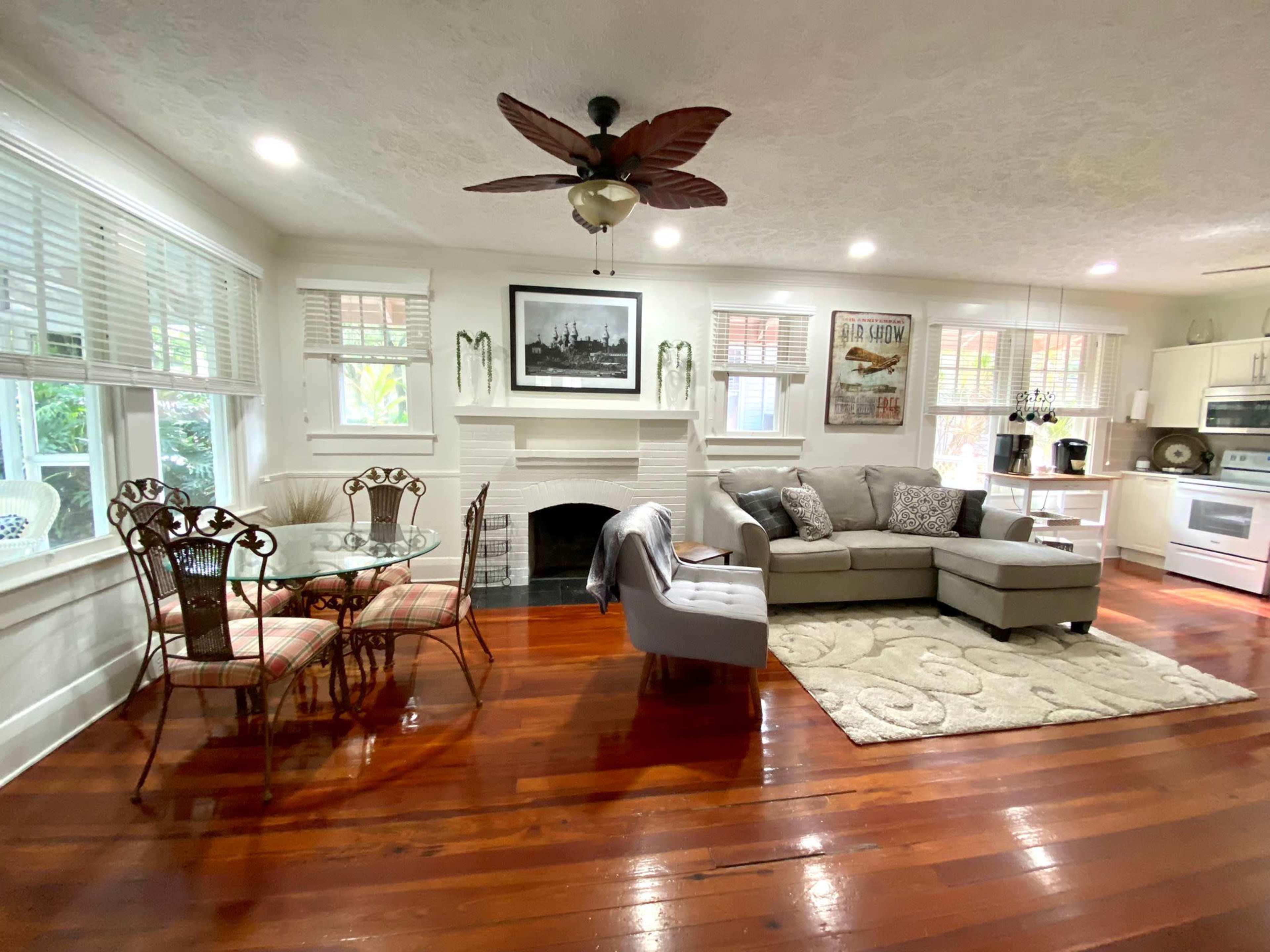 The image shows a cozy living area with a sectional sofa, a coffee table, a dining set, and a fireplace, all featuring polished wooden floors and bright windows.