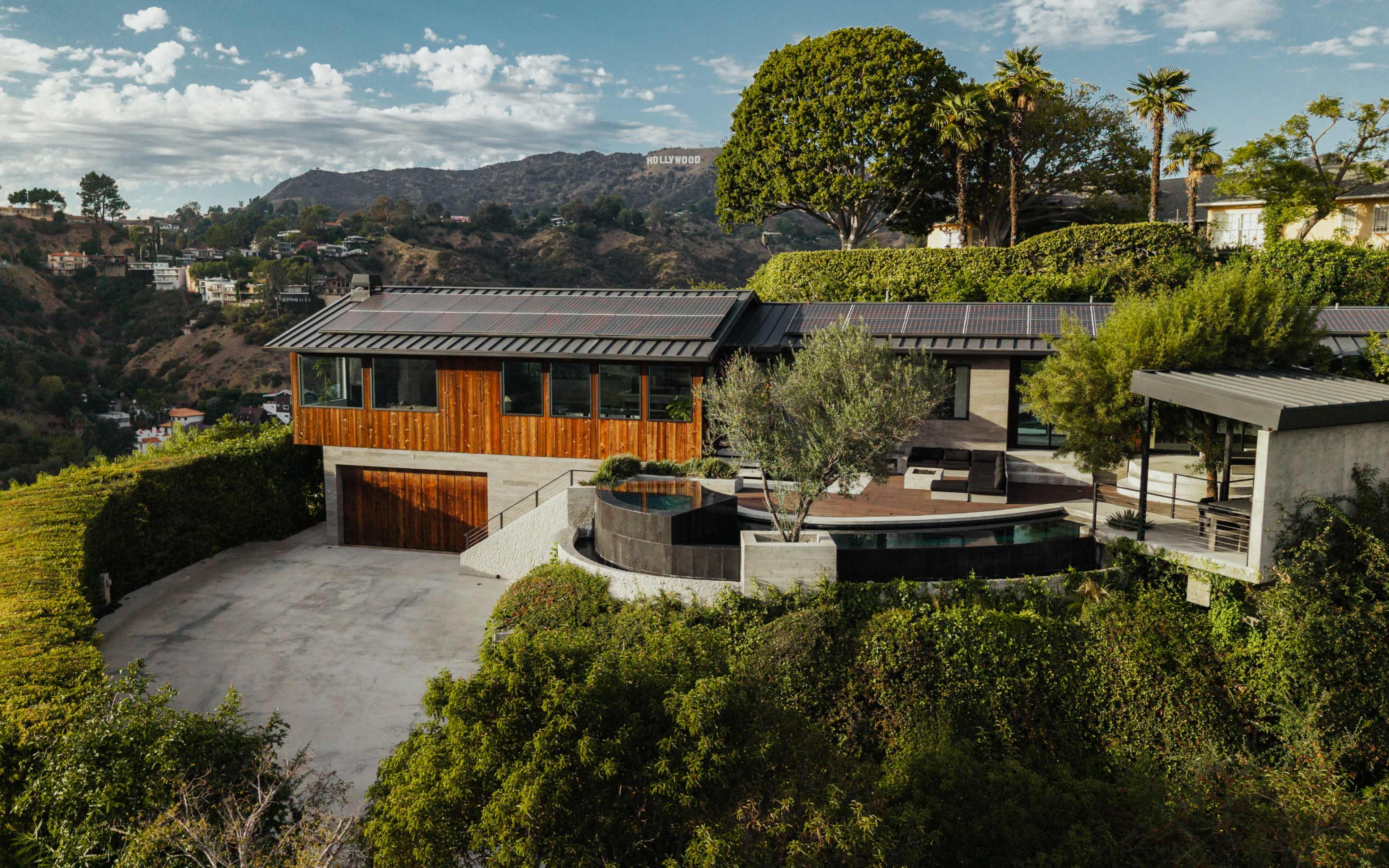 A modern house with a wooden exterior and solar panels is situated on a hillside, surrounded by lush greenery and offering a view of the Hollywood sign in the distance.