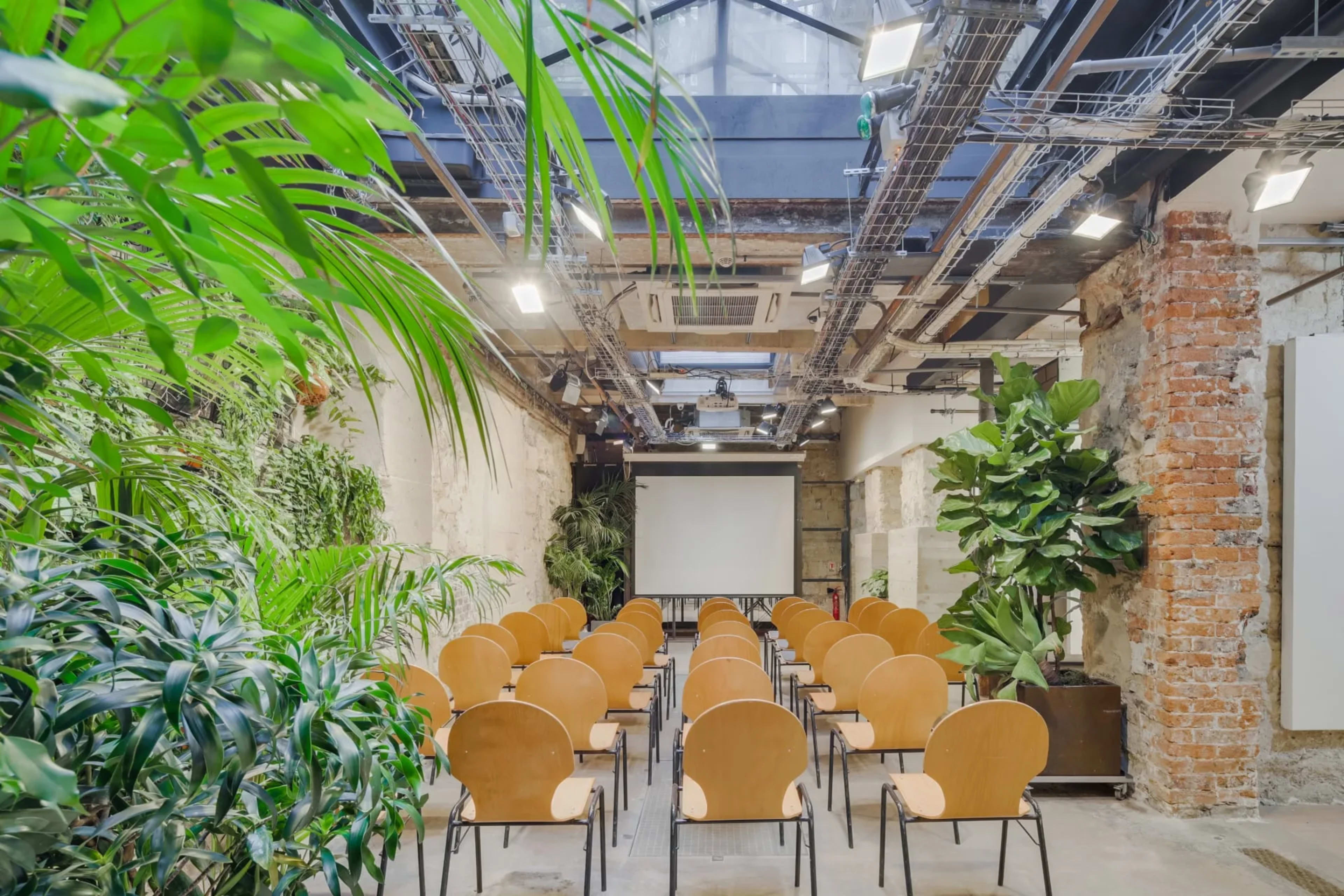 A spacious room with rows of wooden chairs facing a blank projection screen, surrounded by indoor plants and industrial-style decor.