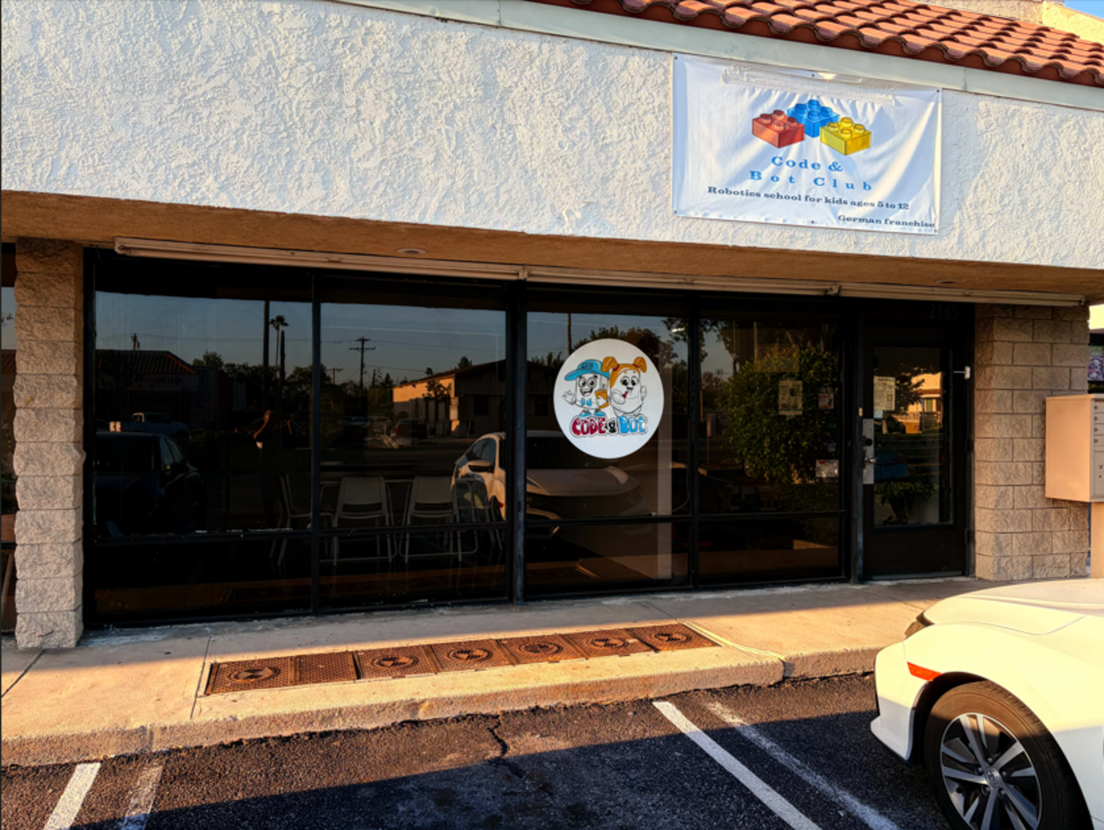 The image shows the exterior of a storefront with large glass windows, a banner above displaying the name "Code & Blox," and a logo on the door featuring cartoon characters.