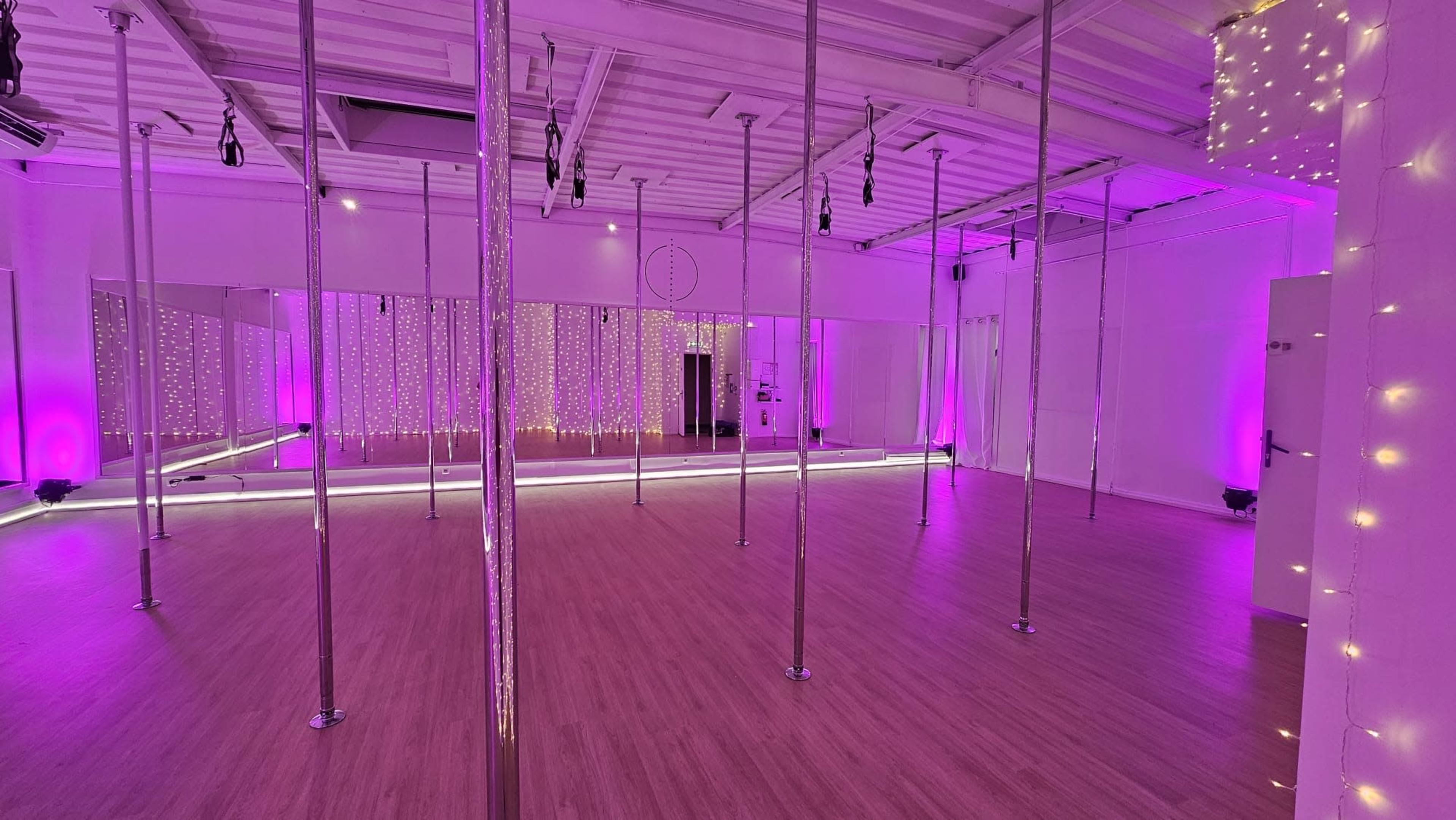 A brightly lit dance studio with polished wooden floors and several vertical poles, accented by purple lighting and decorative string lights on the walls.