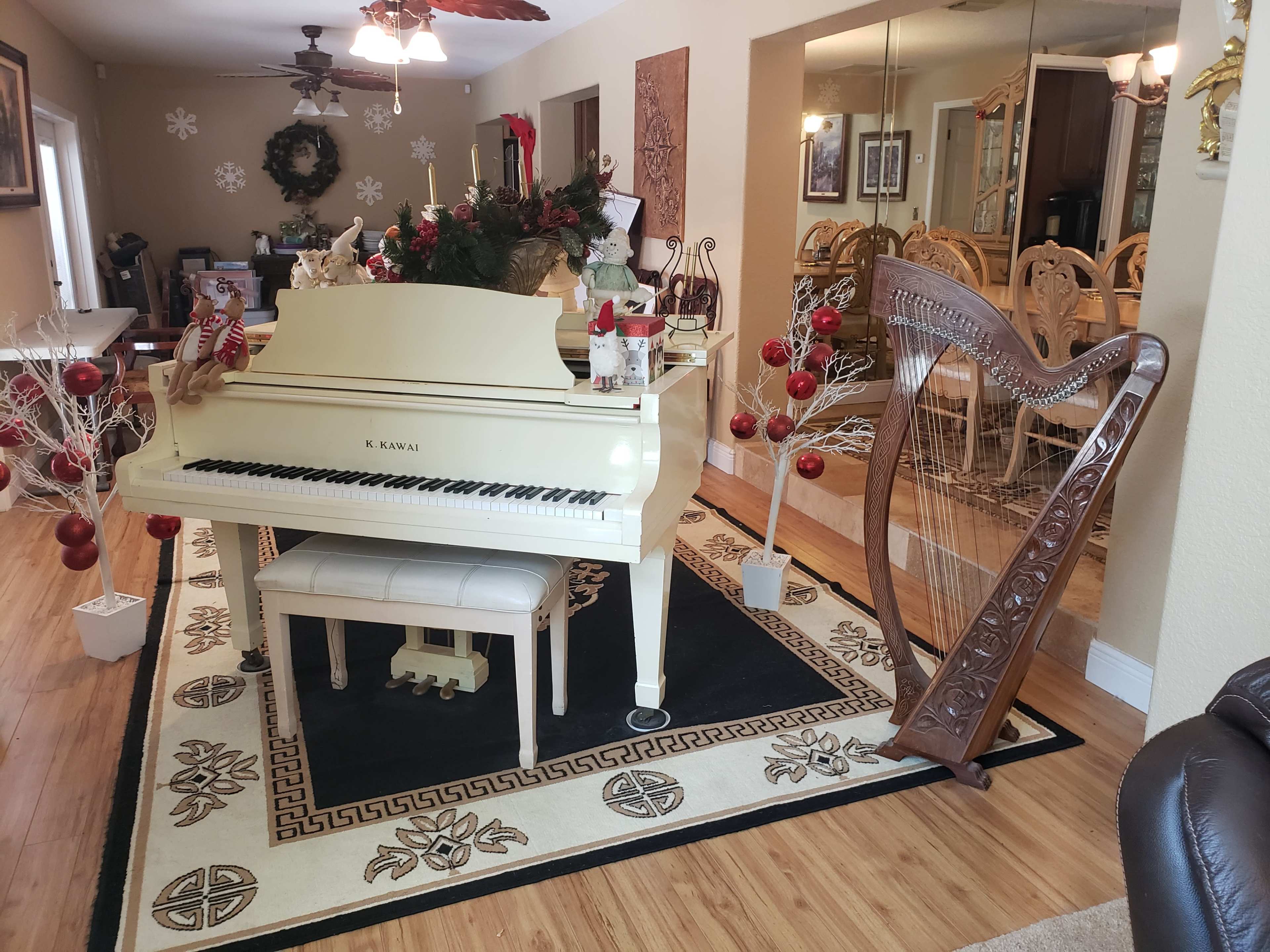 A white piano is positioned in a living room decorated with holiday decor, alongside a wooden harp and ornamental trees with red ornaments.