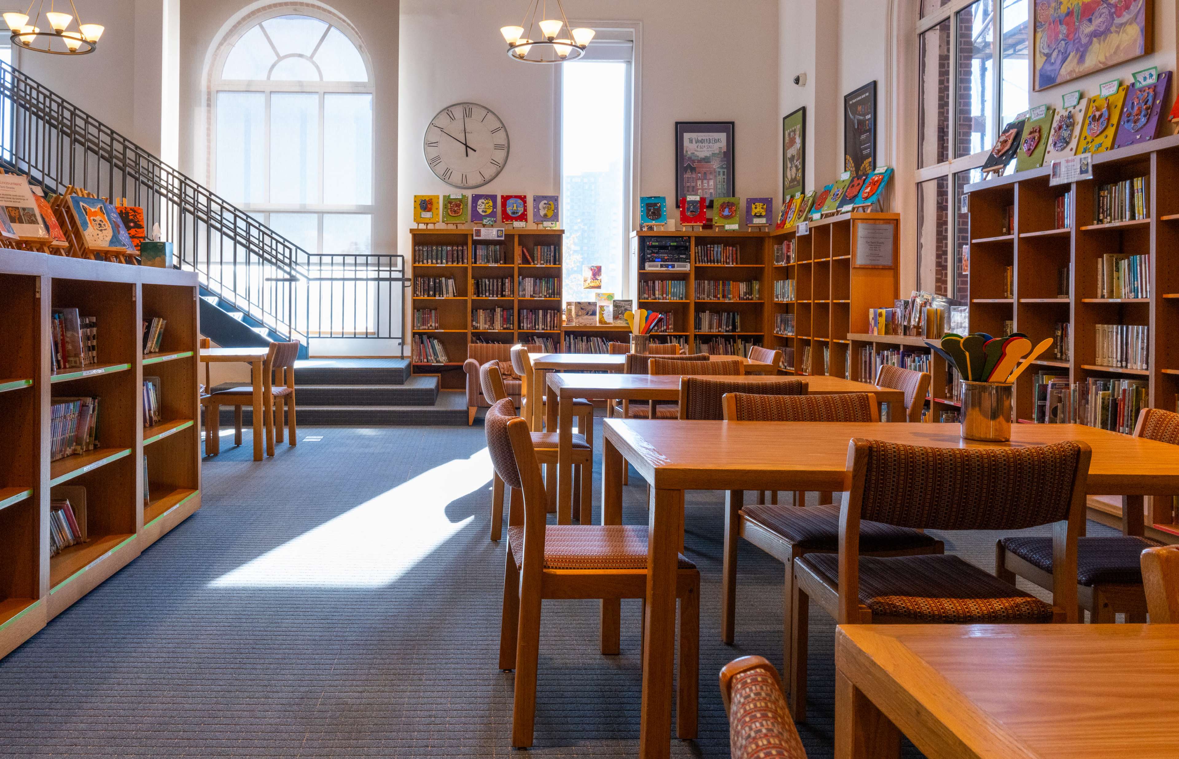A well-lit library features wooden tables and chairs, surrounded by bookshelves filled with colorful books, under large windows and a clock on the wall.