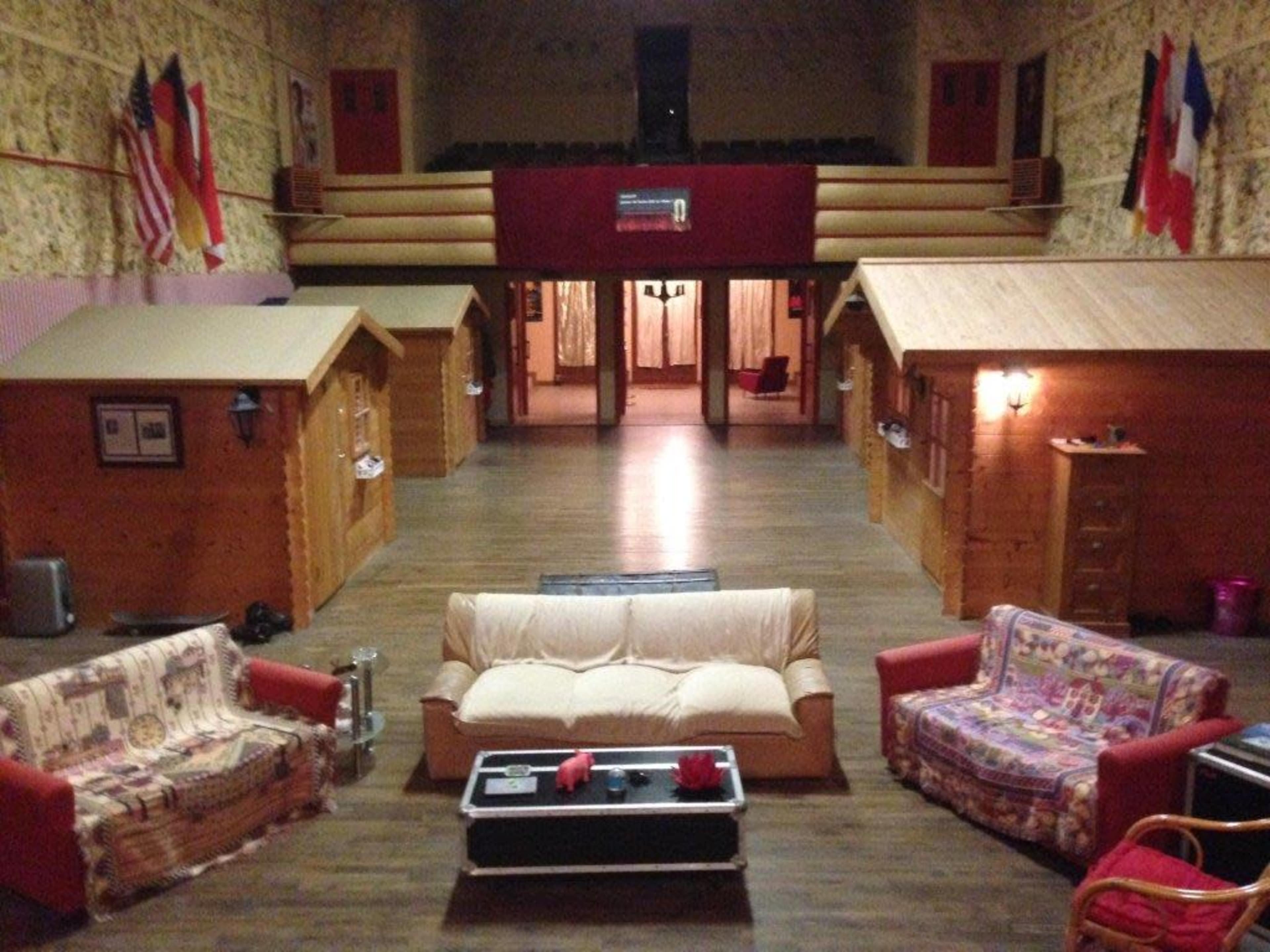 A large room with wooden cabins on either side, two sofas in the foreground, and a stage area at the back with flags hanging on the walls.
