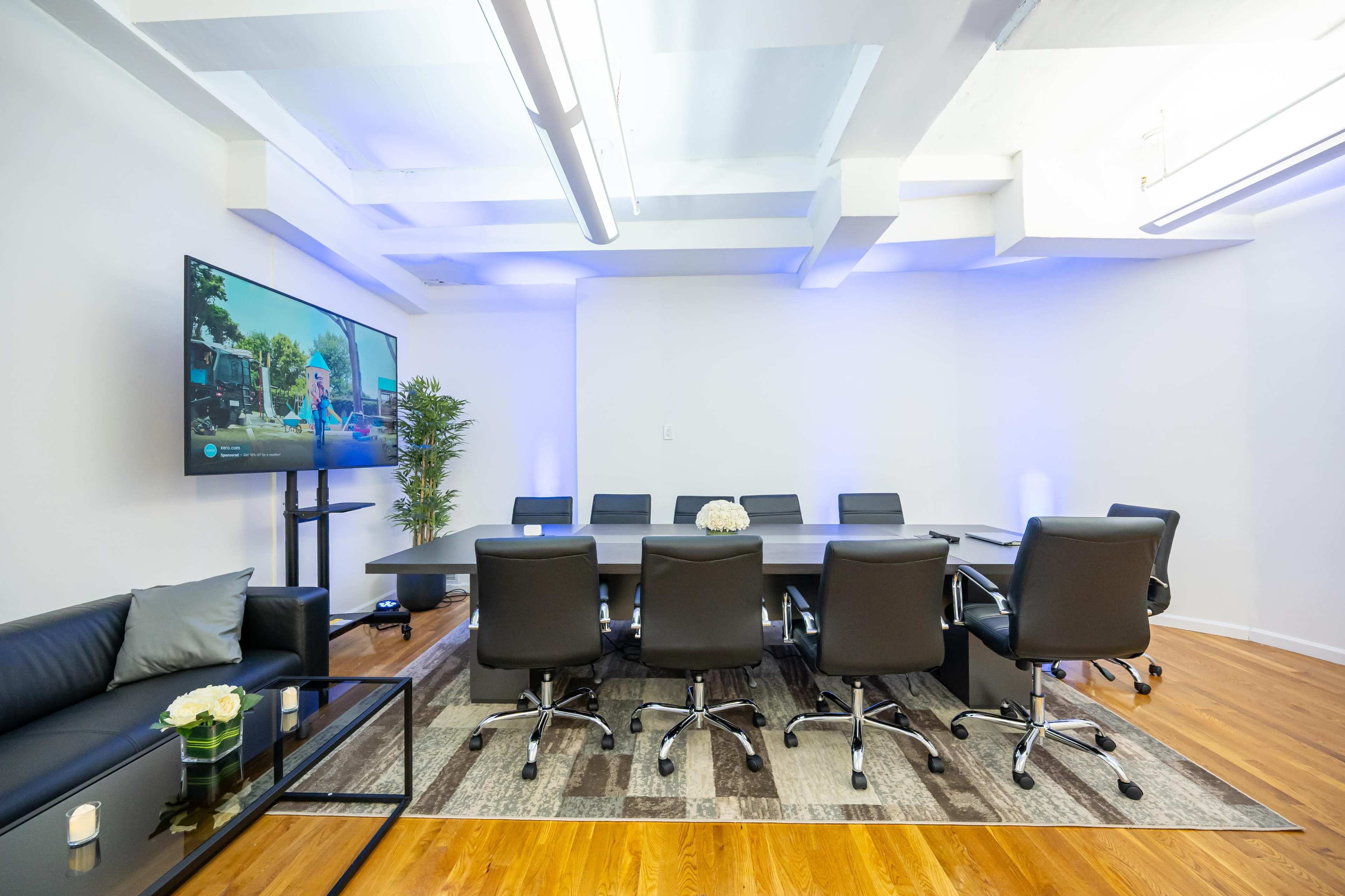A modern conference room features a long table with black chairs, a large television screen, and a sofa with a coffee table.