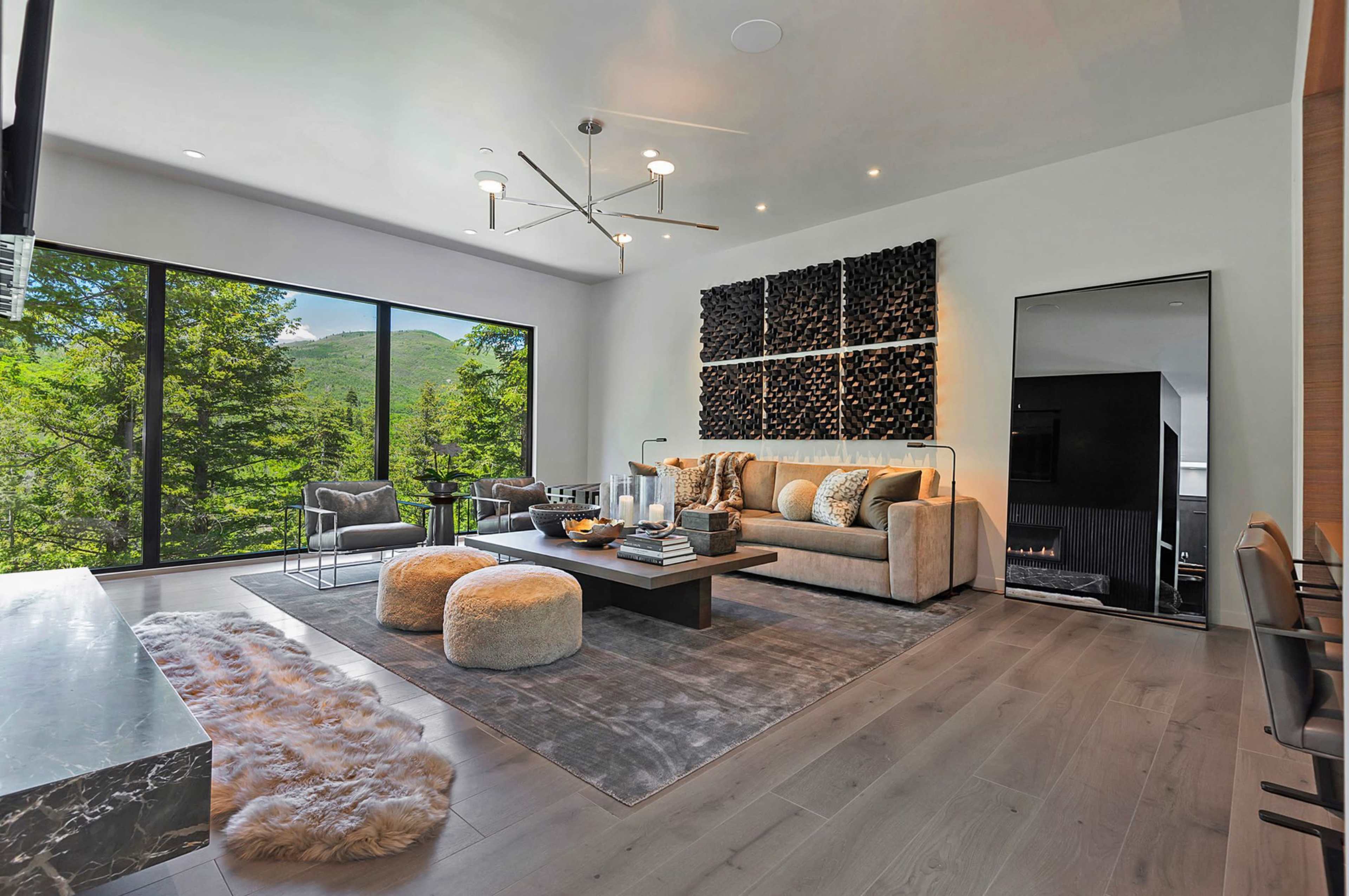 The modern living room features large windows overlooking greenery, a comfortable sofa, a coffee table, and a soft rug on the wooden floor.