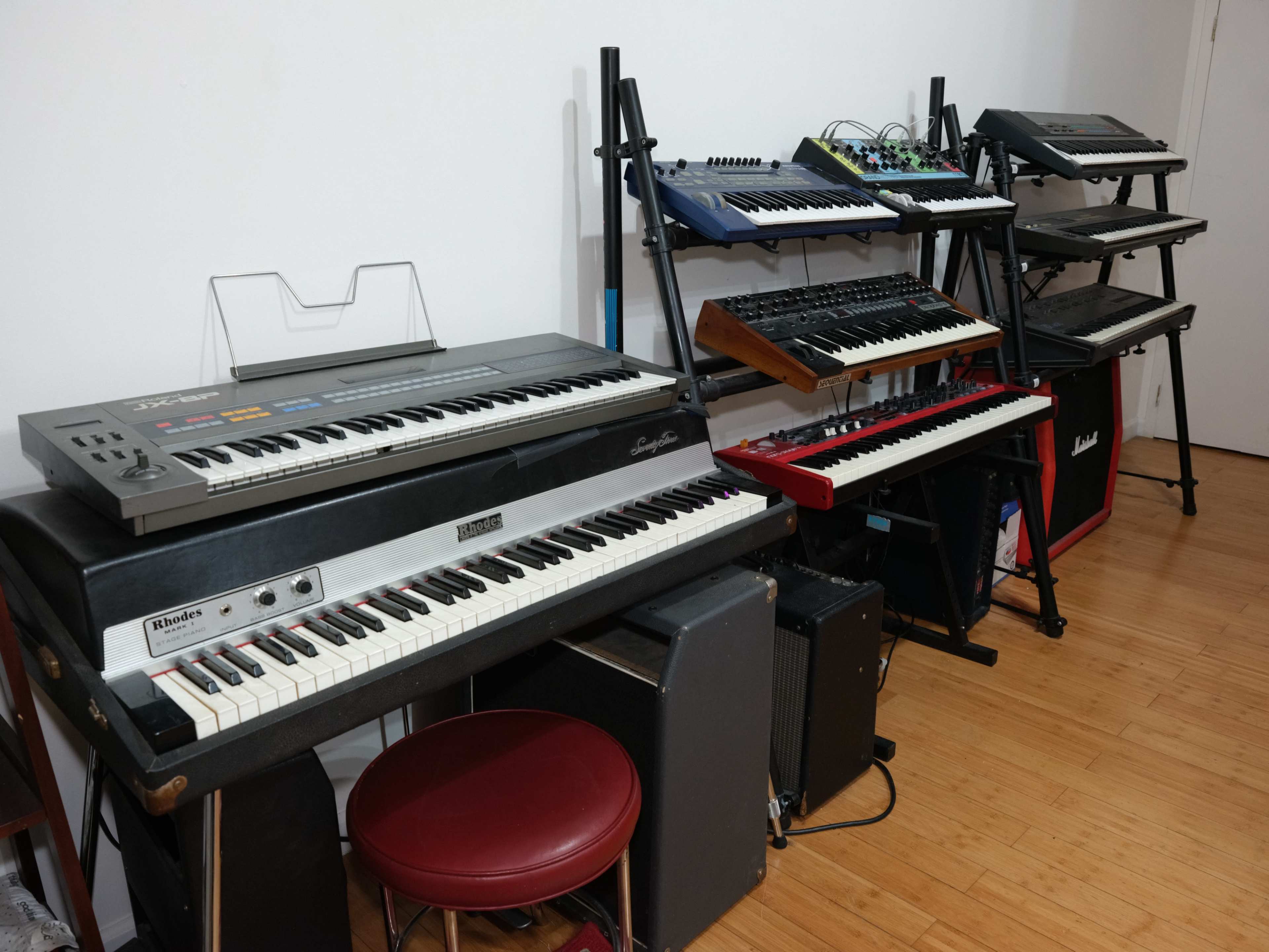 A collection of vintage synthesizers and keyboards is arranged against a wall, with a red keyboard and a stool in the foreground.