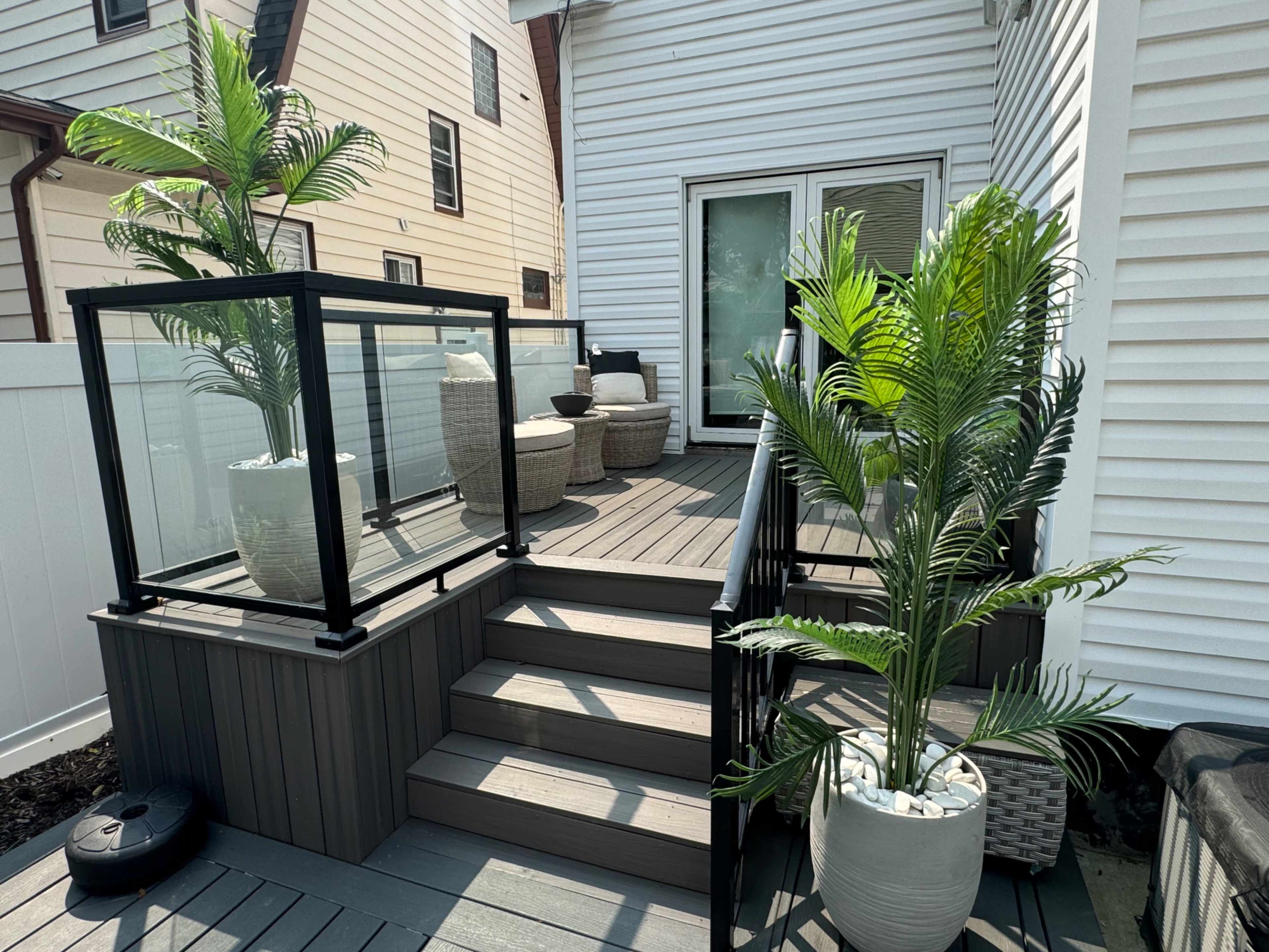 The image shows a wooden deck with stairs, featuring potted plants and a glass railing, adjacent to a house.