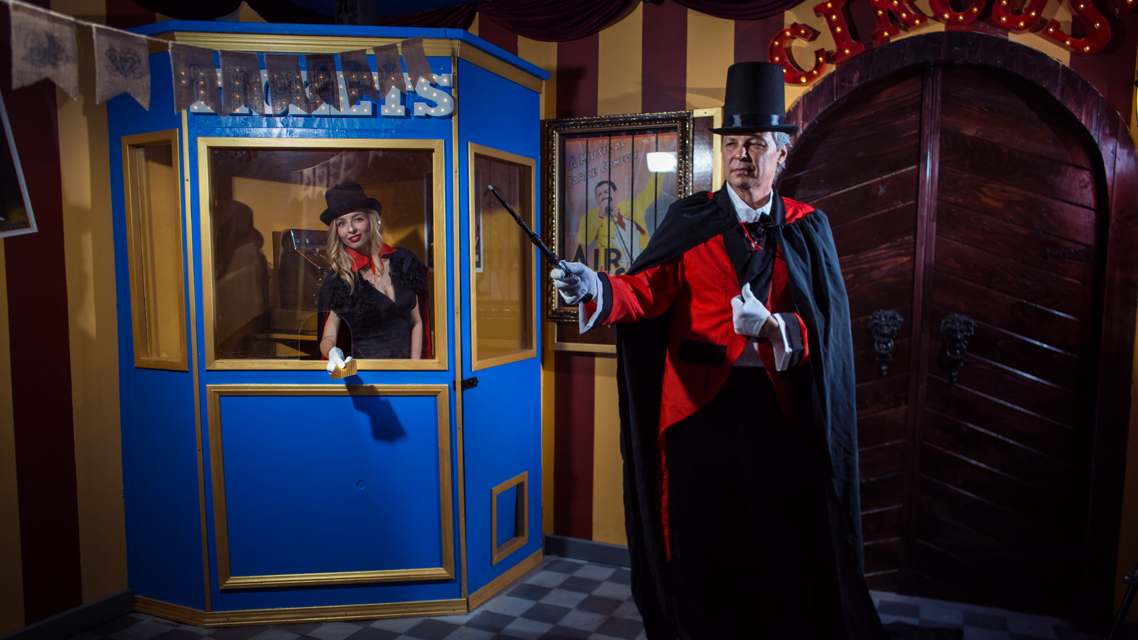 A man in a top hat and a cape holds a wand while standing in front of a ticket booth, where a woman in a black dress smiles at him.