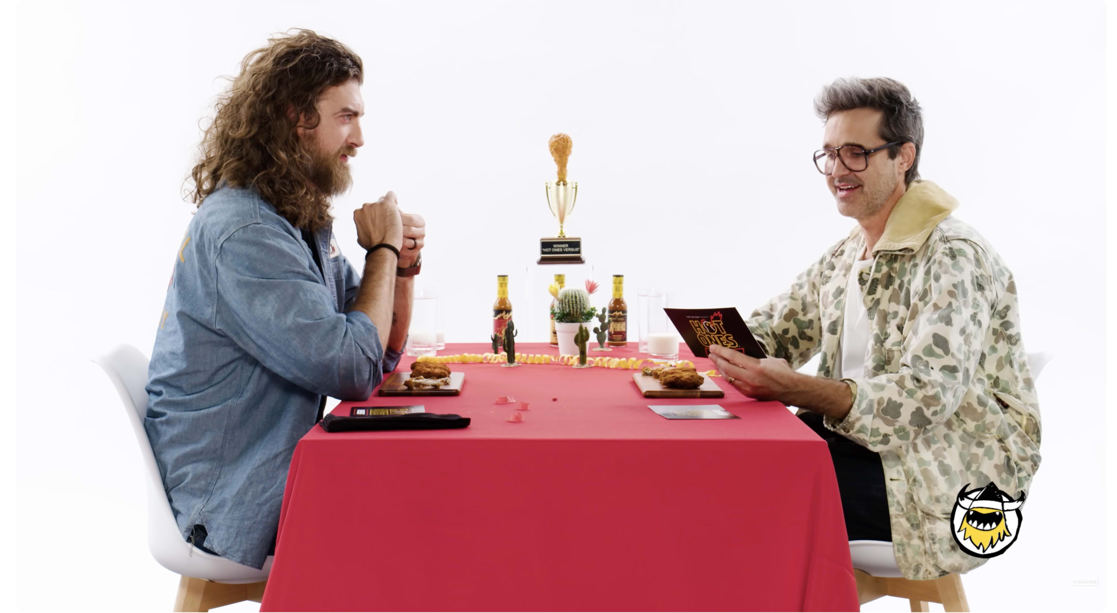 Two men sit at a table covered with a red tablecloth, each holding a menu, with food plates in front of them and a trophy displayed between them.