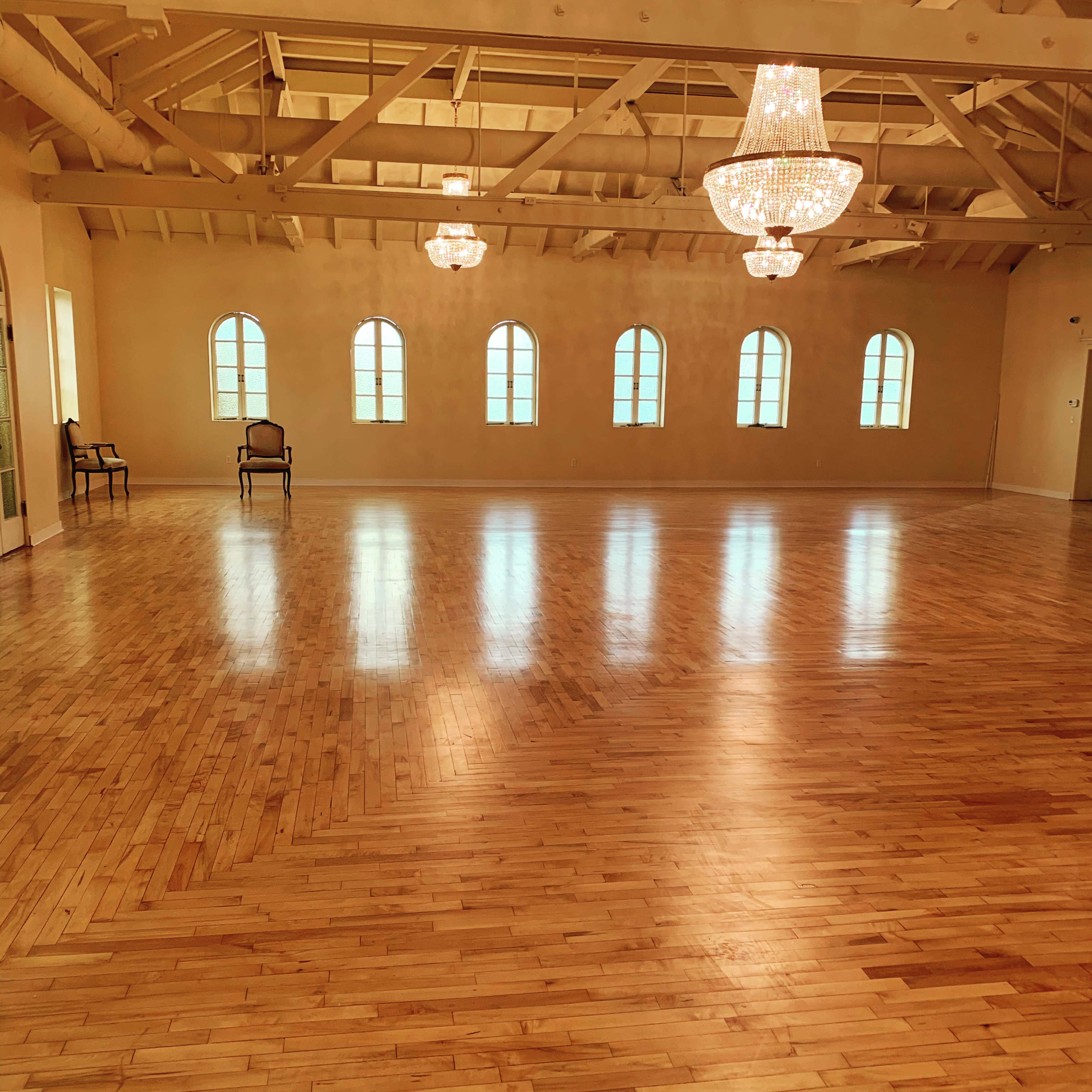 A spacious dance studio with polished wooden floors, large windows, and elegant chandeliers.