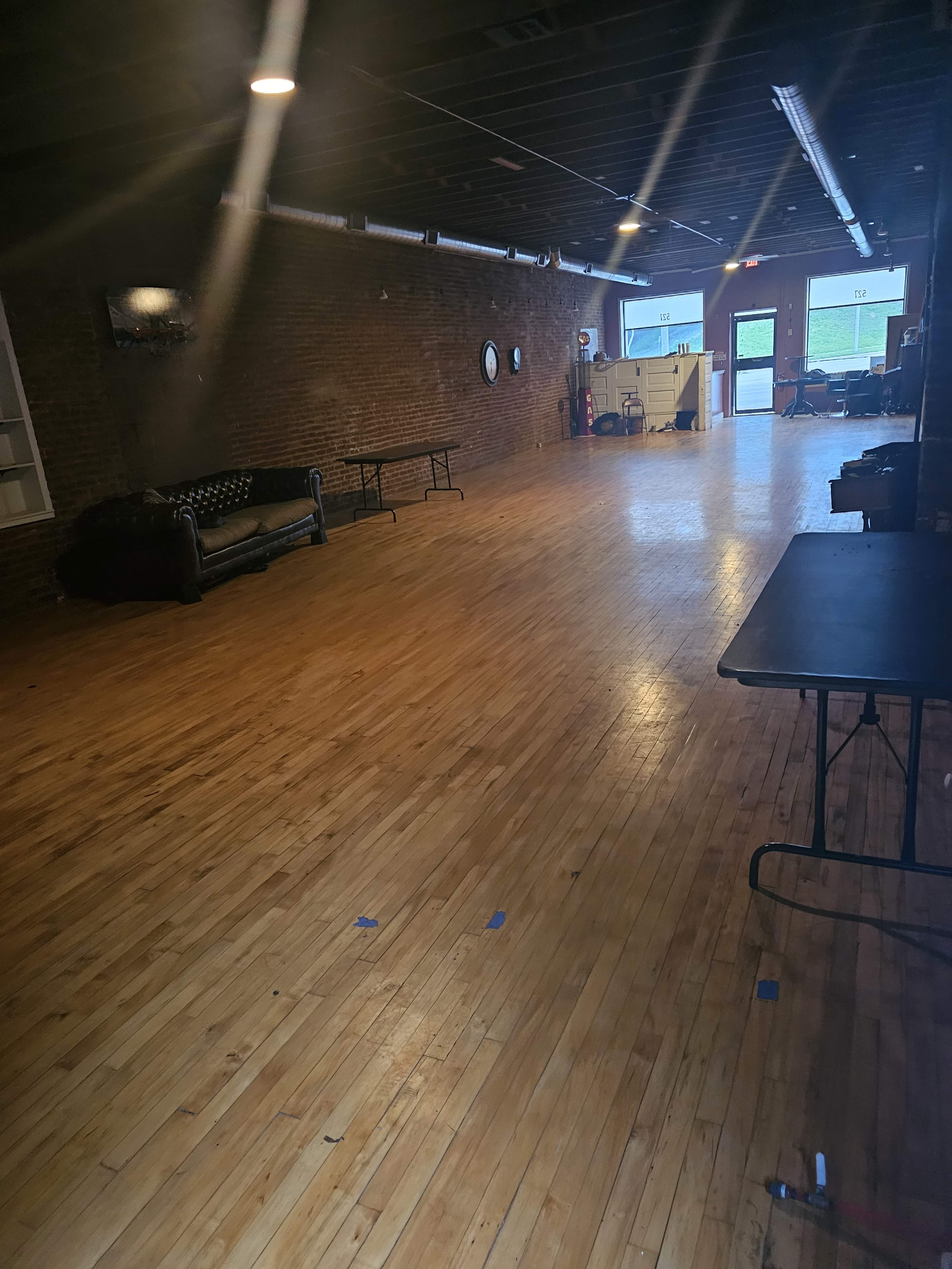 An empty room with wooden flooring, exposed brick walls, and a few pieces of furniture, including a couch and tables.