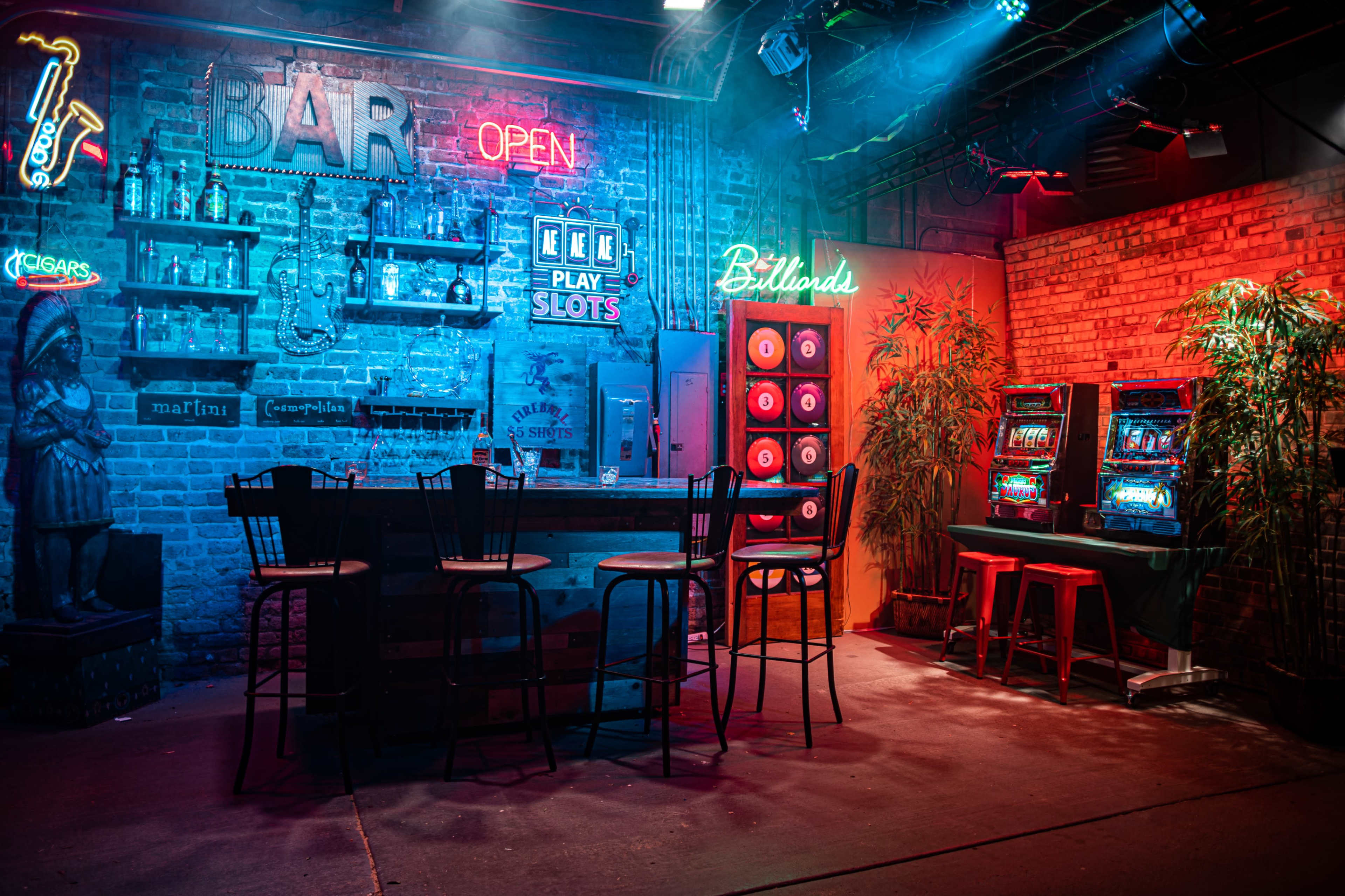 A dimly lit bar setting with neon signs, a wooden bar counter, and several bar stools, alongside two slot machines and a brick wall adorned with a billiards area.