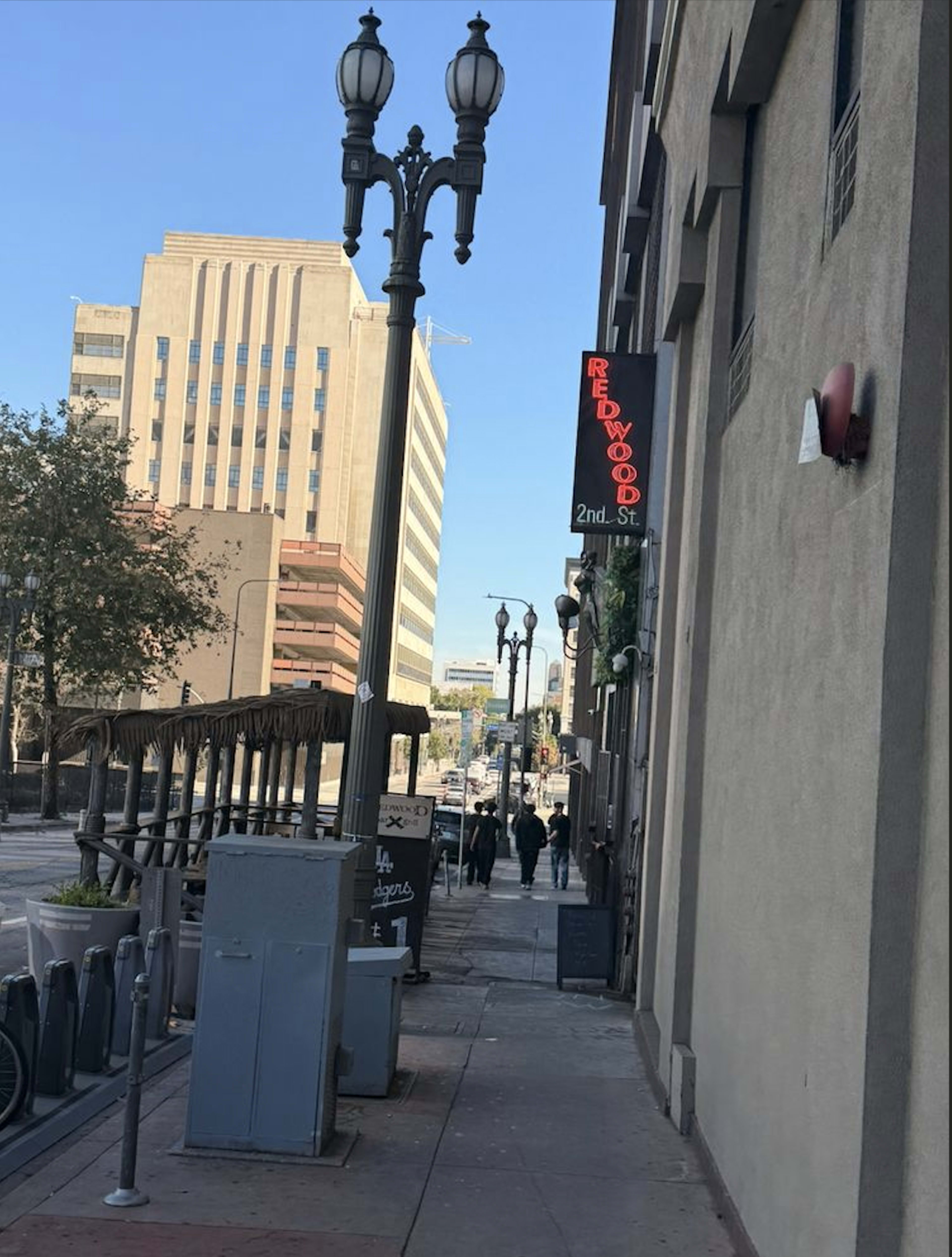 The image shows a city street with a restaurant sign for "Redwood" on a building, street lamps lining the sidewalk, and pedestrians walking in the distance.