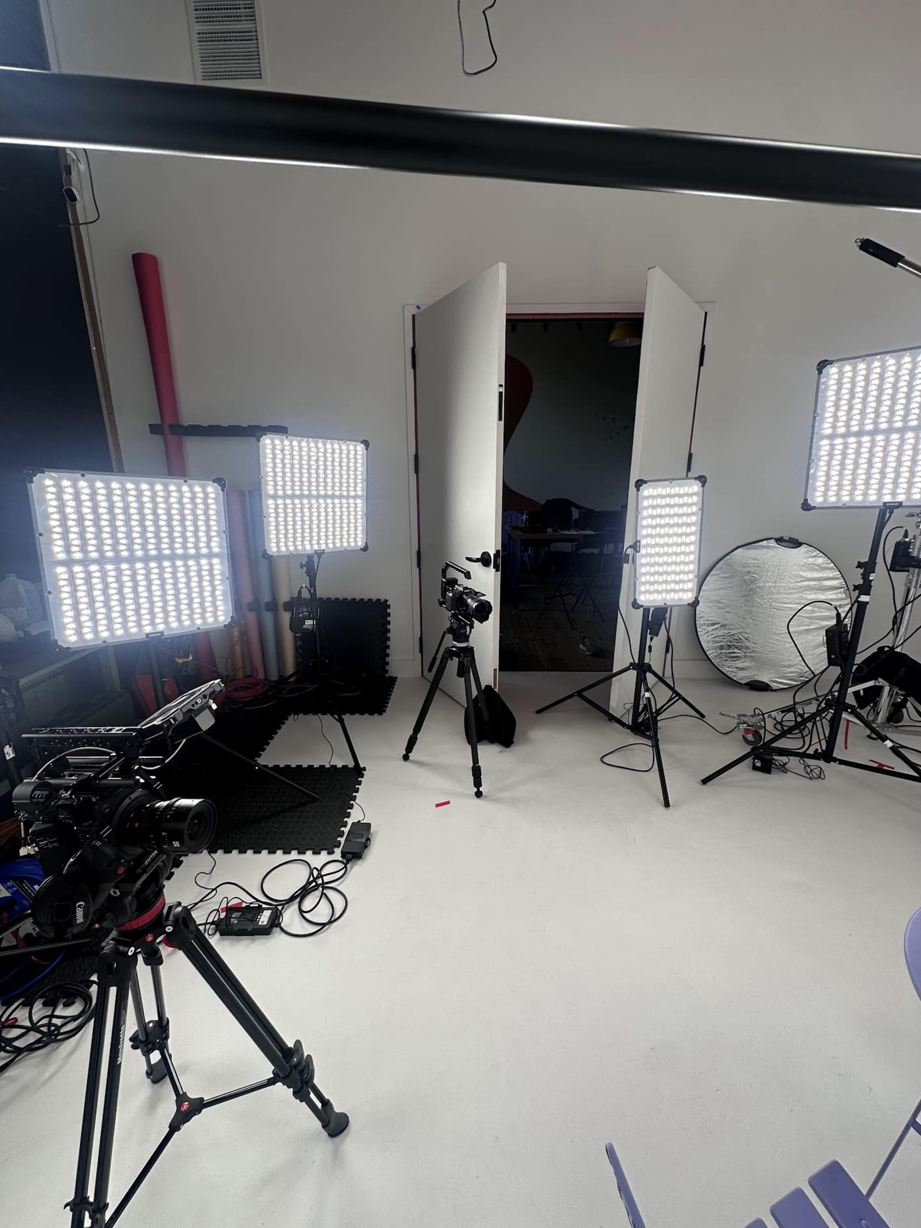 The image shows a photo studio setup with multiple lights, cameras on tripods, and a backdrop area.