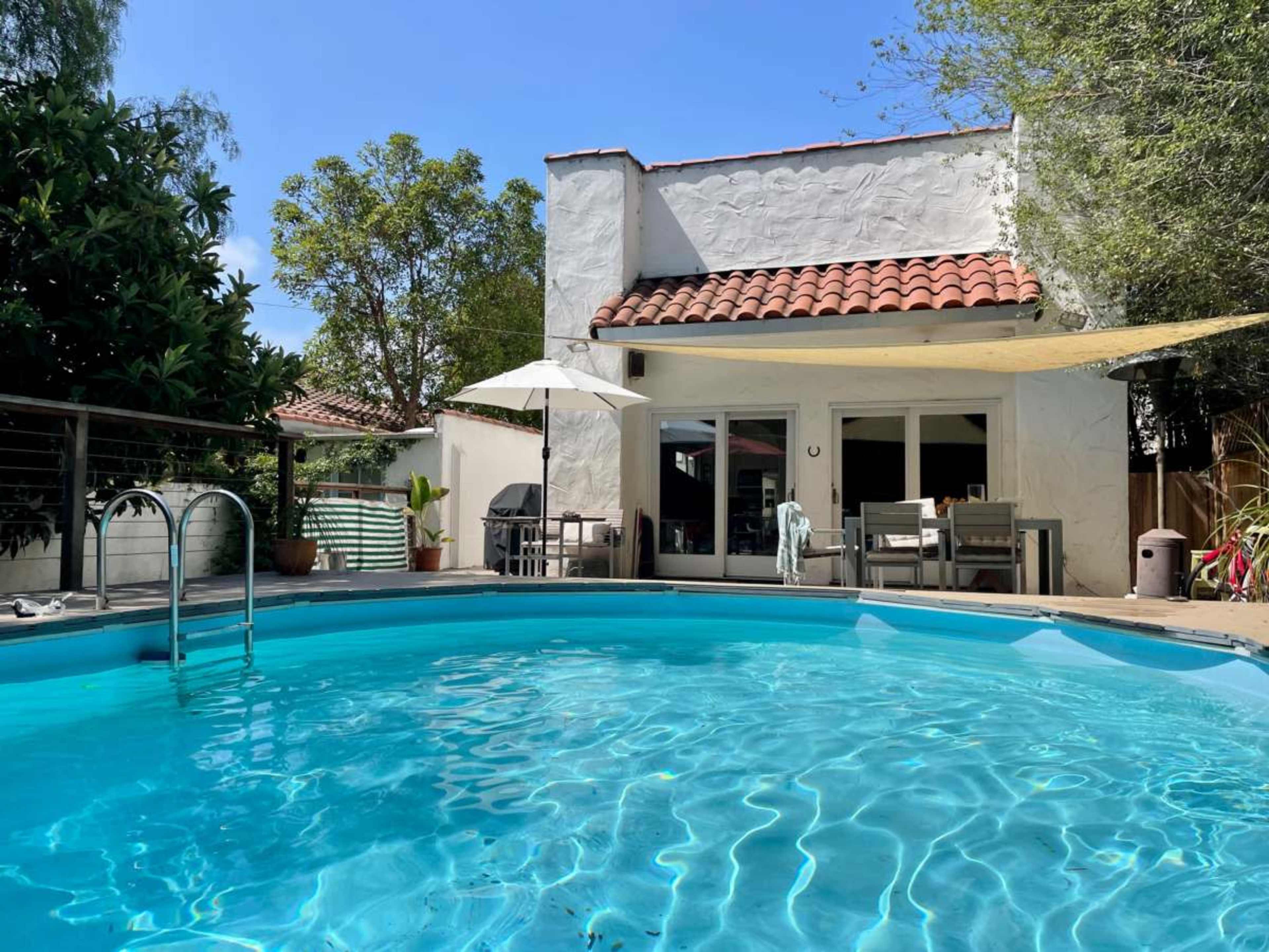 A round swimming pool reflects the clear blue sky next to a modern house with a tiled roof and outdoor furniture.