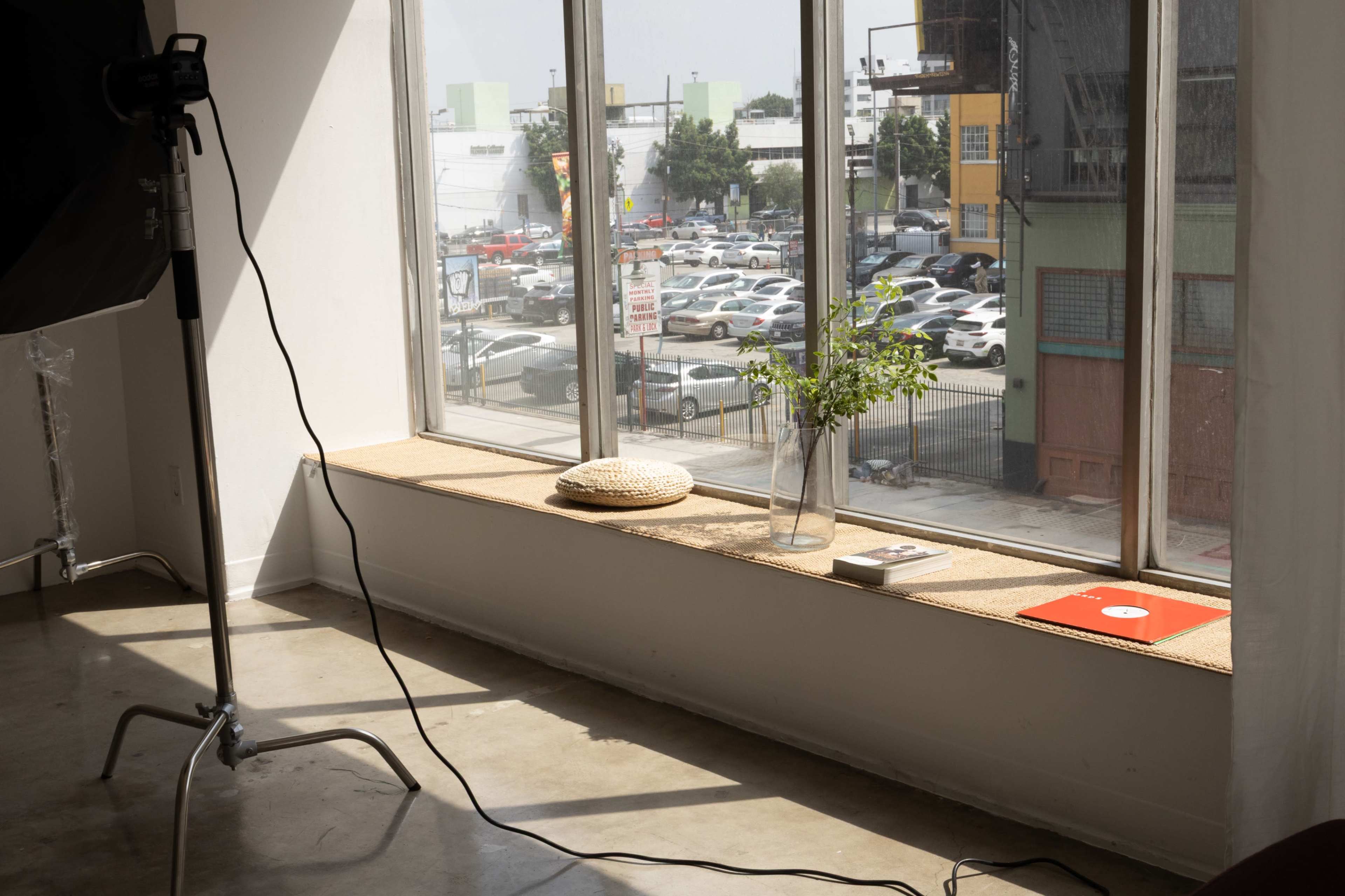 A bright, spacious room features a large window with a view of an outdoor parking area, and a windowsill displaying a vase with greenery, a round cushion, and a book.