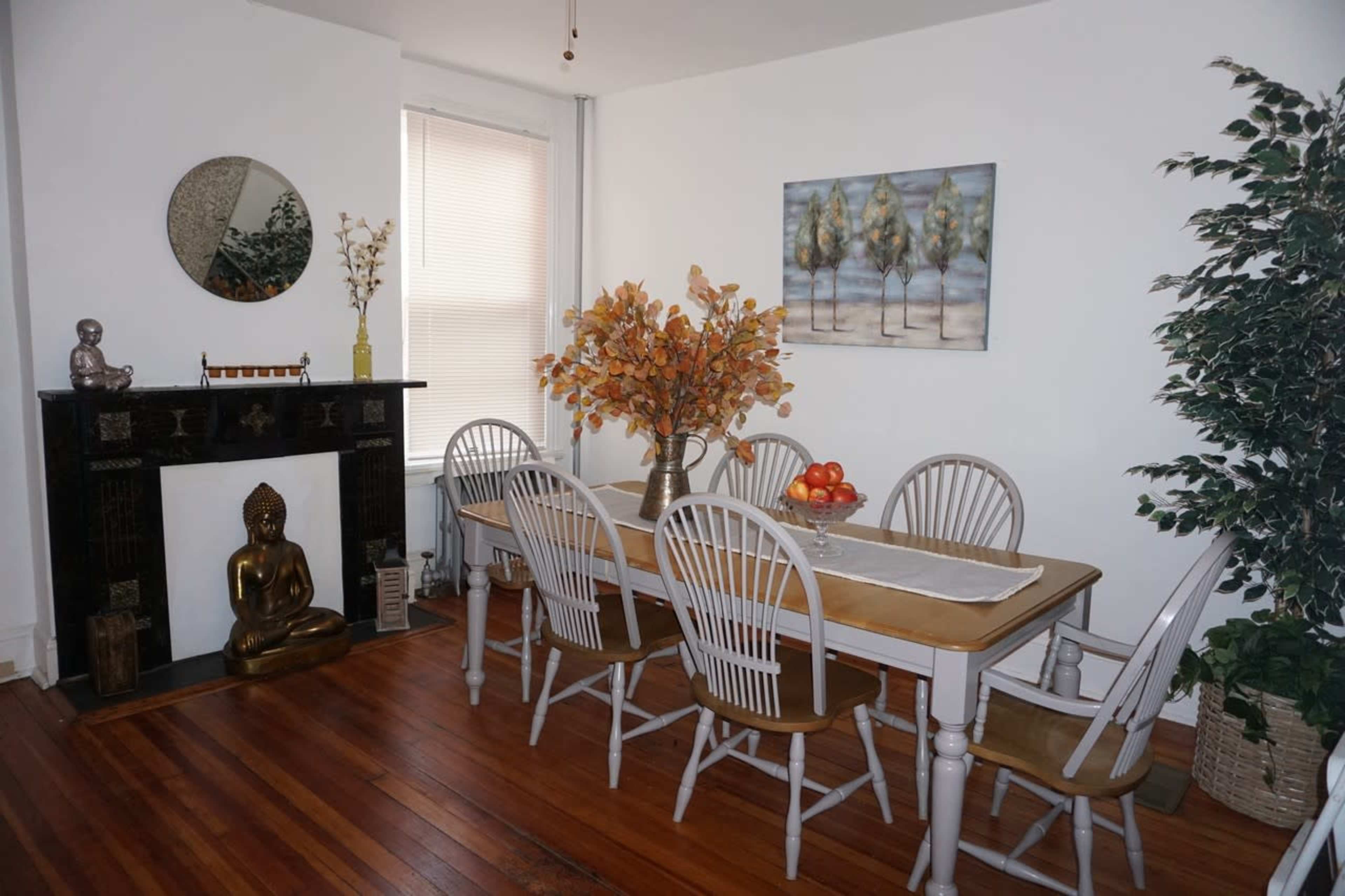 A dining room features a long table surrounded by wooden chairs, with a decorative centerpiece of autumn foliage and a statue of a seated figure on a mantel.