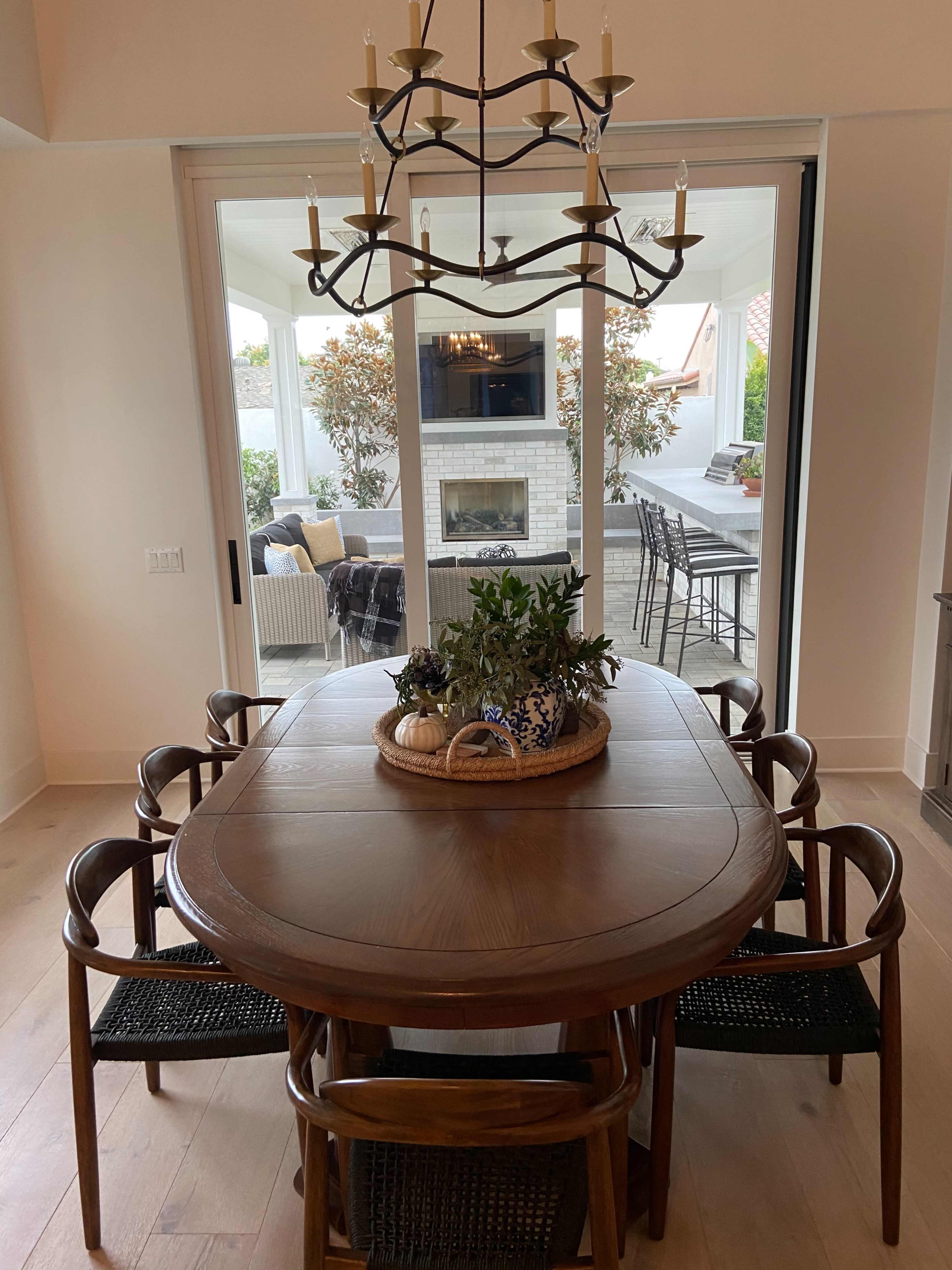 A wooden dining table with a central plant arrangement is surrounded by six chairs, and large glass doors lead to a patio area with outdoor seating and a fireplace.