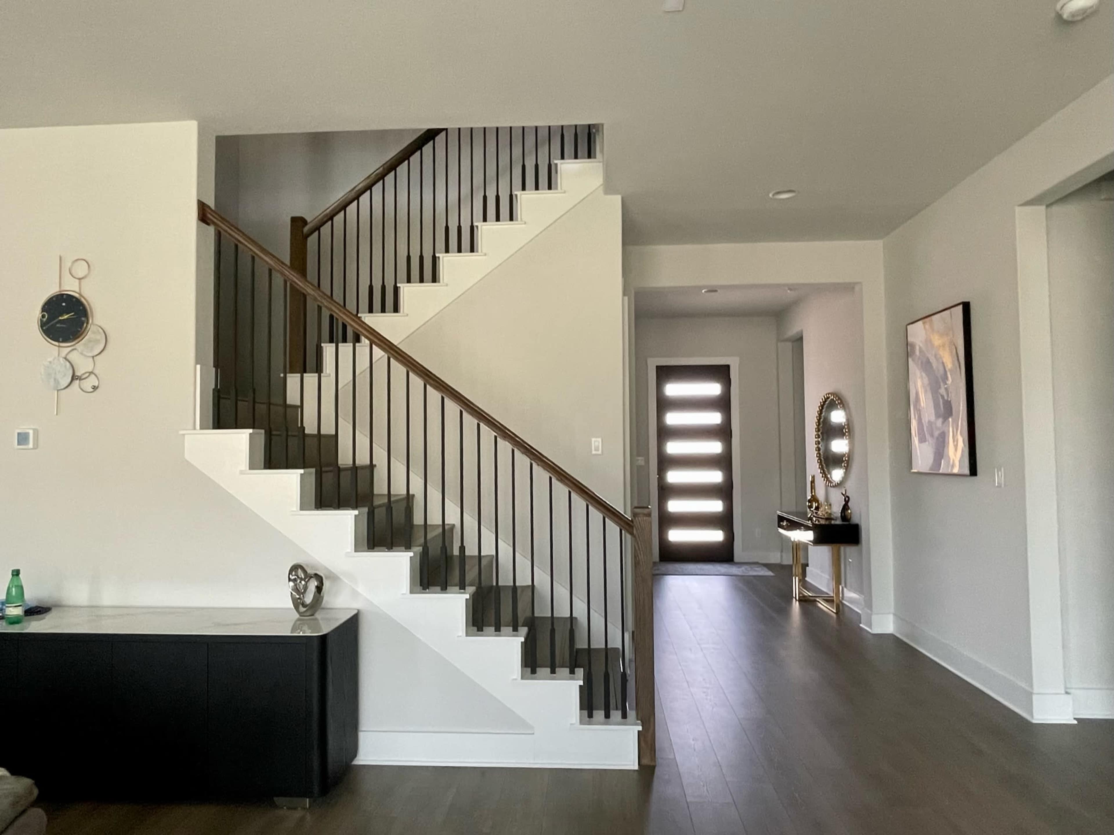 The image shows a modern entryway with a staircase leading to an upper level, a front door with multiple panels, and a decorative side table with a mirror.