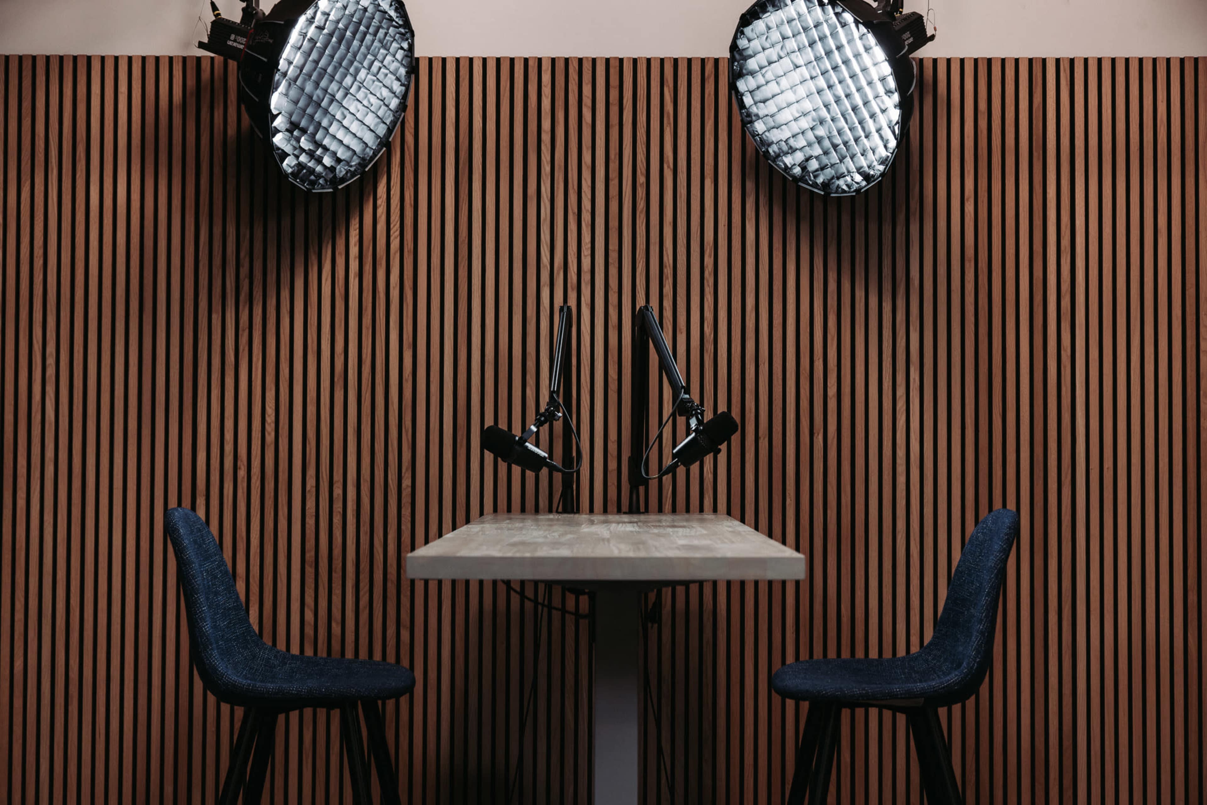 A table with two chairs is positioned under bright studio lights against a wall of vertical wooden slats.