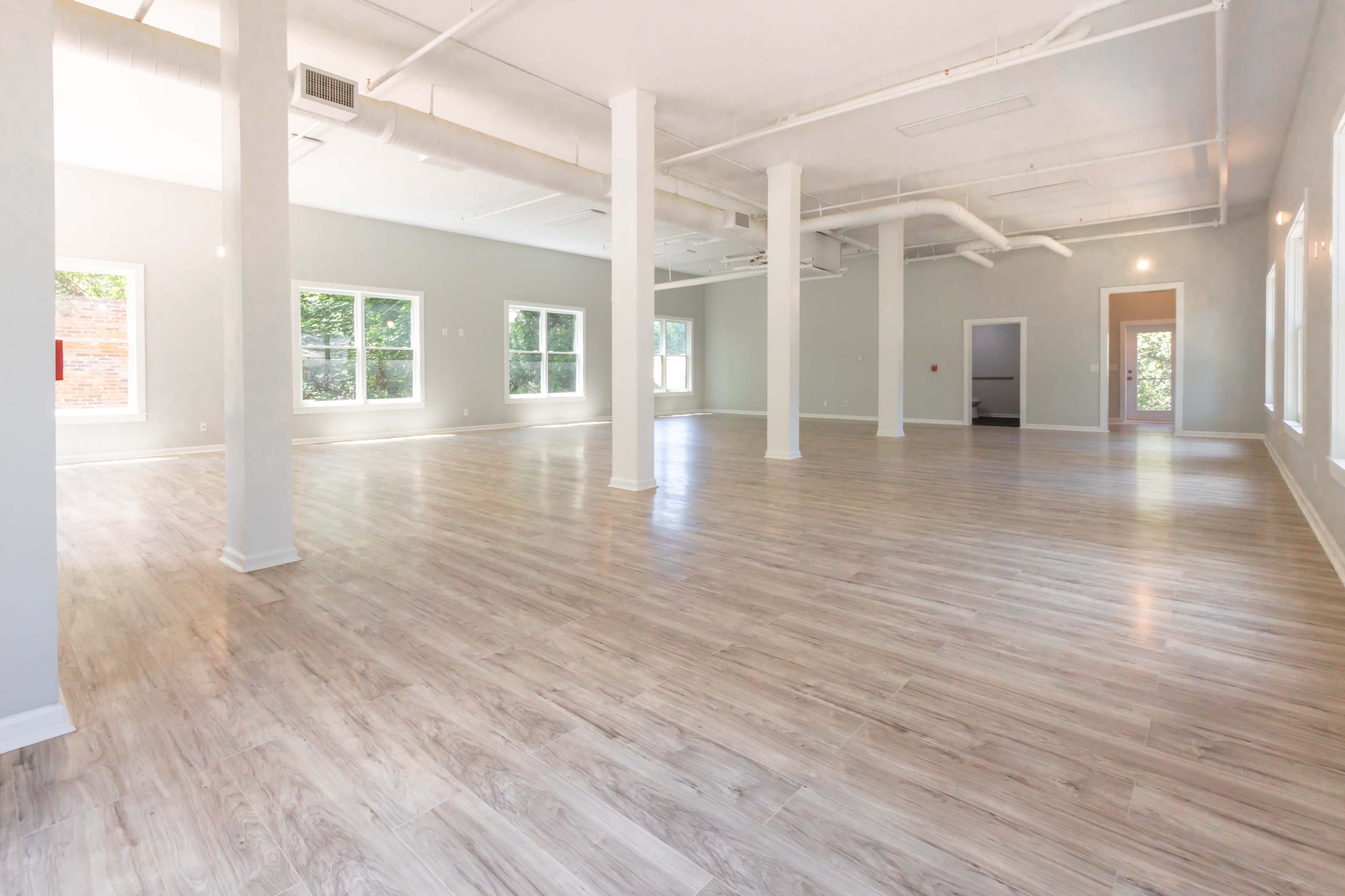The image shows a spacious, empty room with wooden flooring, large windows, and light-colored walls.