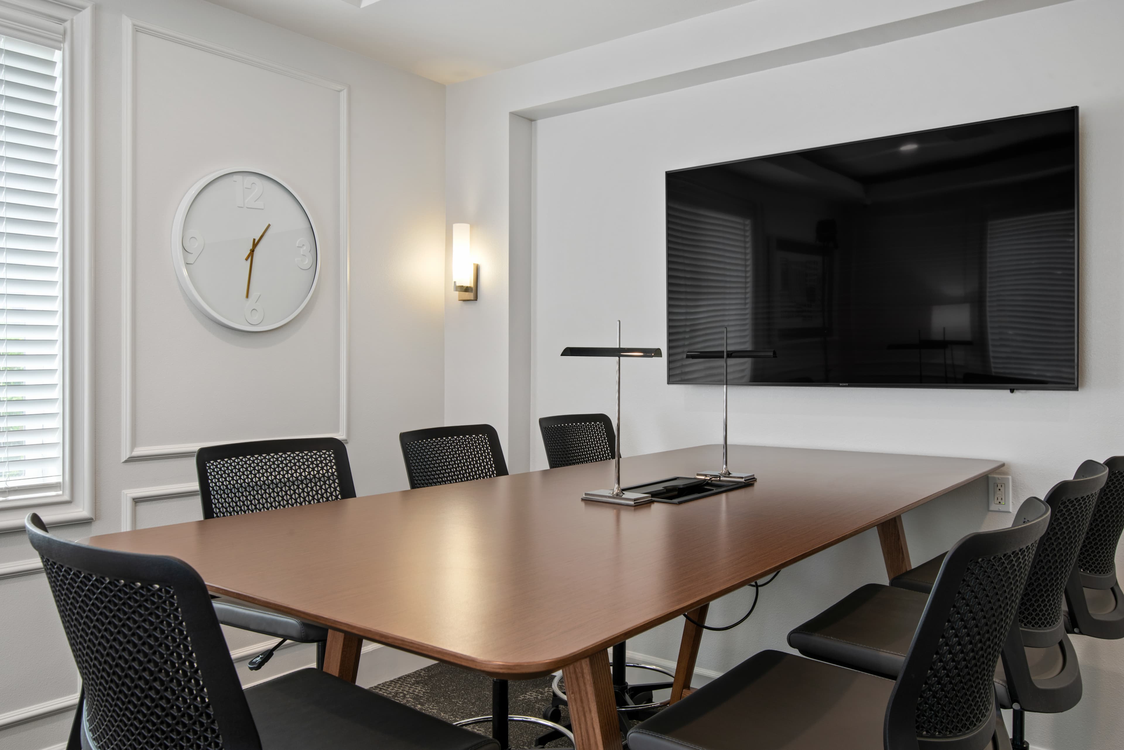 A modern conference room features a large wooden table surrounded by black mesh chairs, a mounted flat-screen TV, and a wall clock.