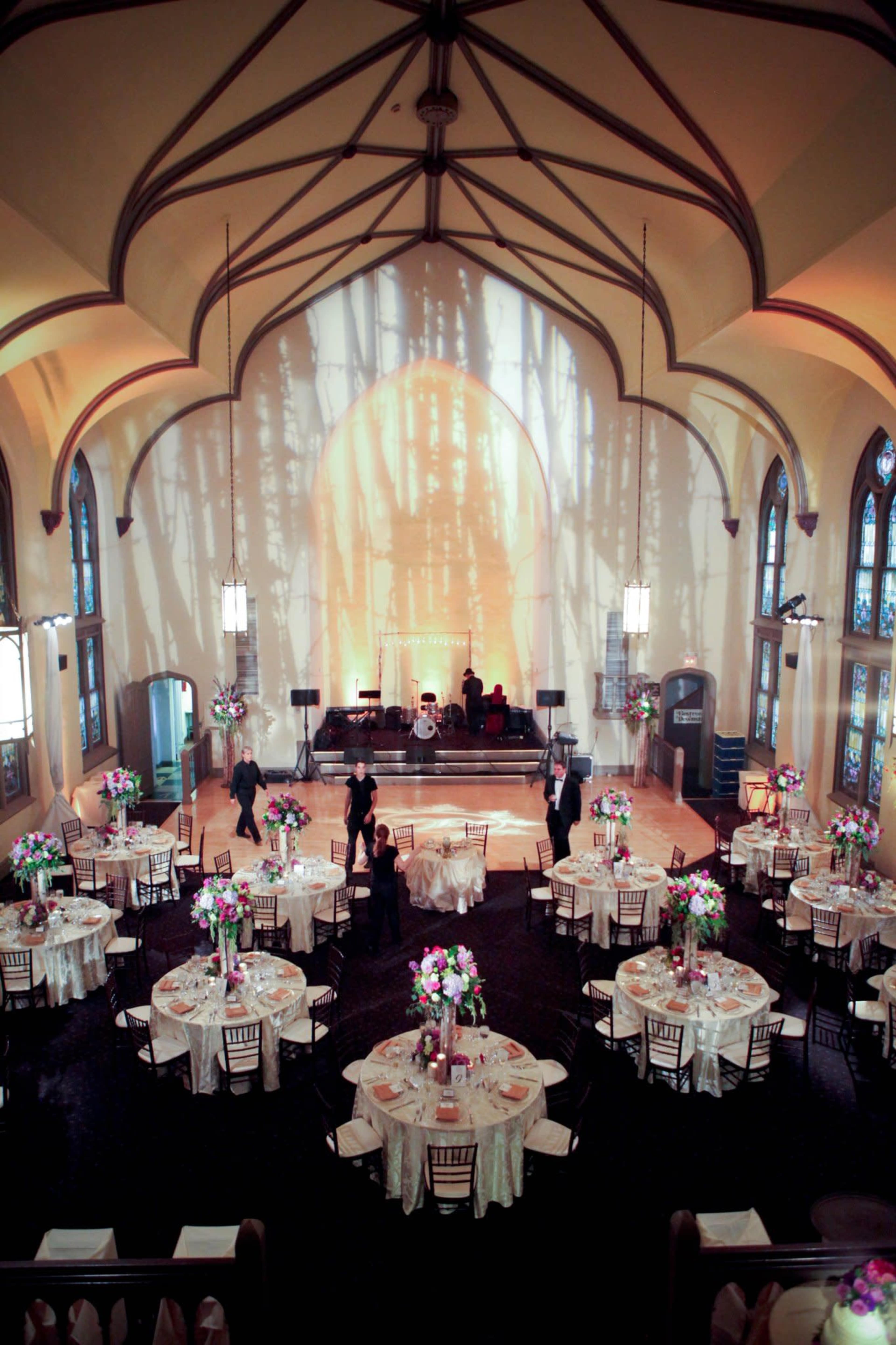 A beautifully arranged event space featuring round tables with floral centerpieces, set against a backdrop of a stage with instruments in a venue with high arched ceilings and large windows.
