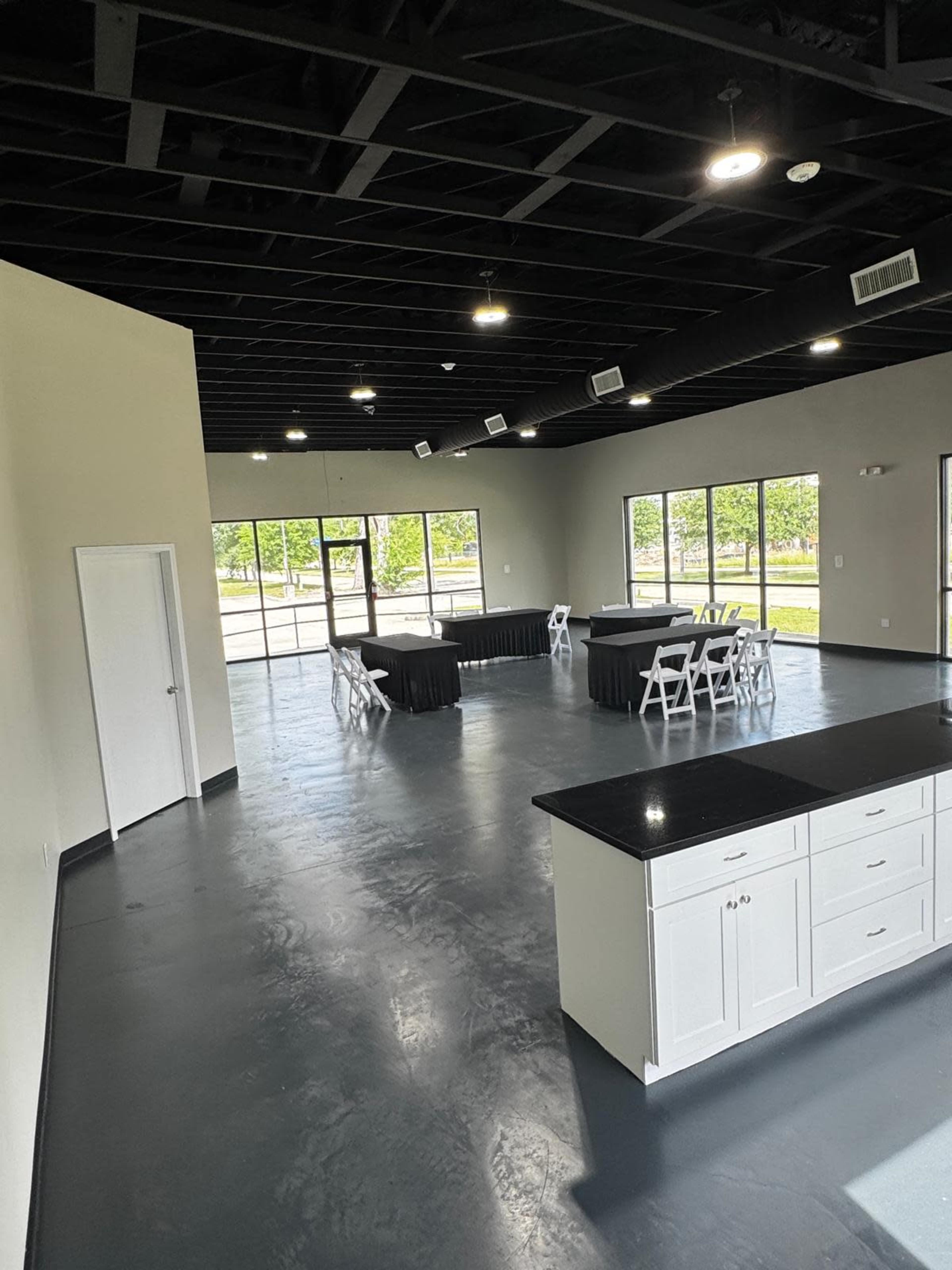The image shows a spacious, well-lit room with black tables accompanied by white chairs, and large windows letting in natural light.