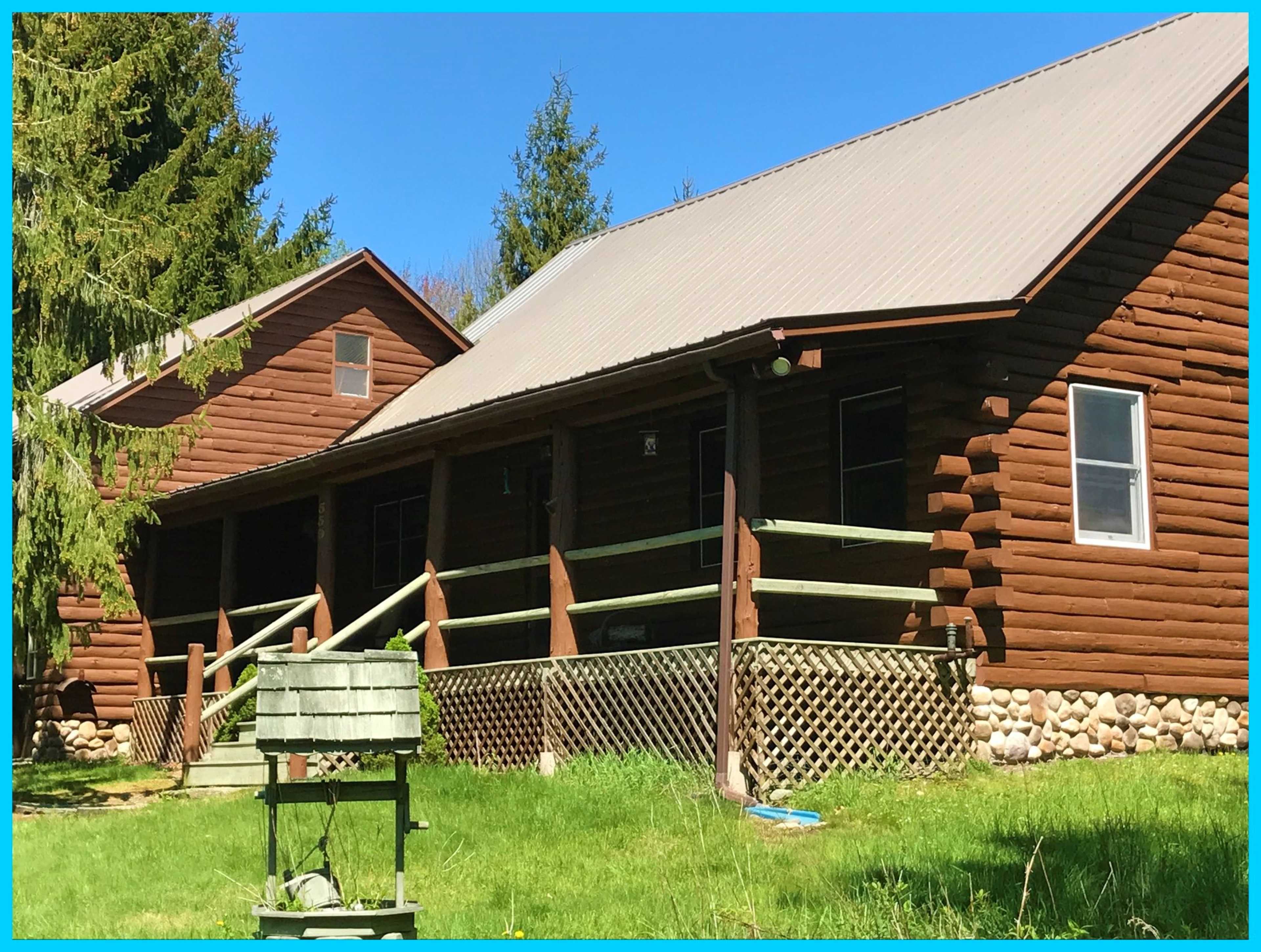 A log cabin with a metal roof sits in a grassy area, featuring a front porch and stone foundation.