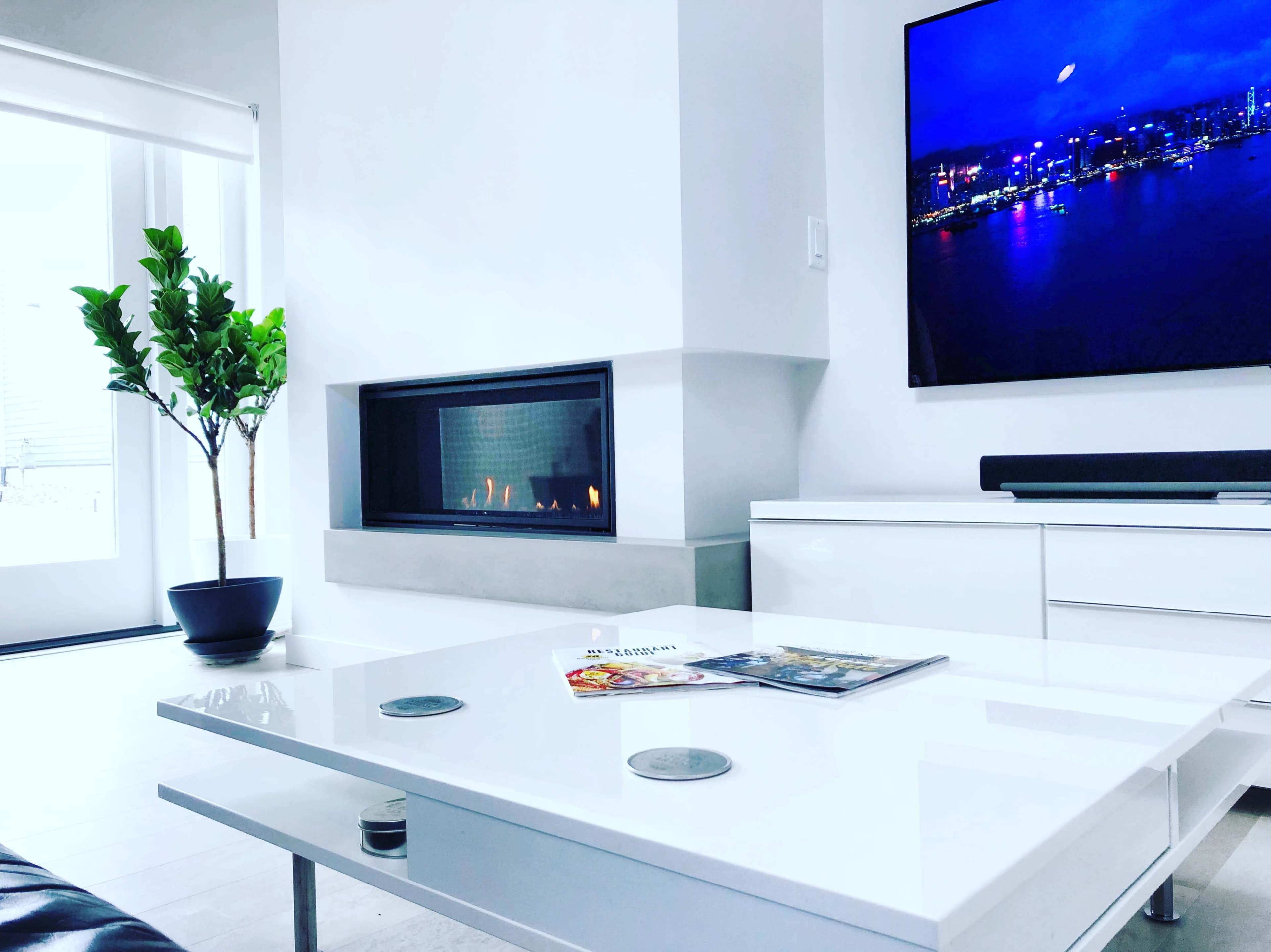 A modern living room with a sleek white coffee table, a flat-screen television mounted on the wall, a fireplace, and a potted plant in the corner.