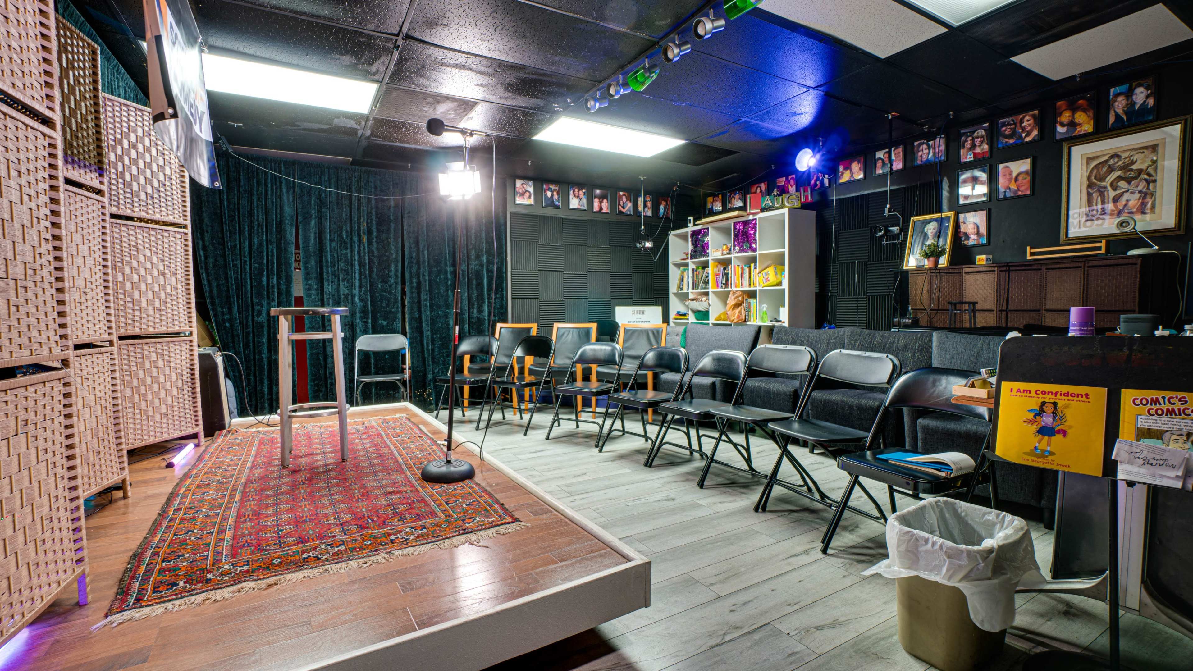 The image shows a small stage area with a microphone and a table, surrounded by a semicircle of black chairs, with a backdrop featuring black curtains and framed photographs on the walls.