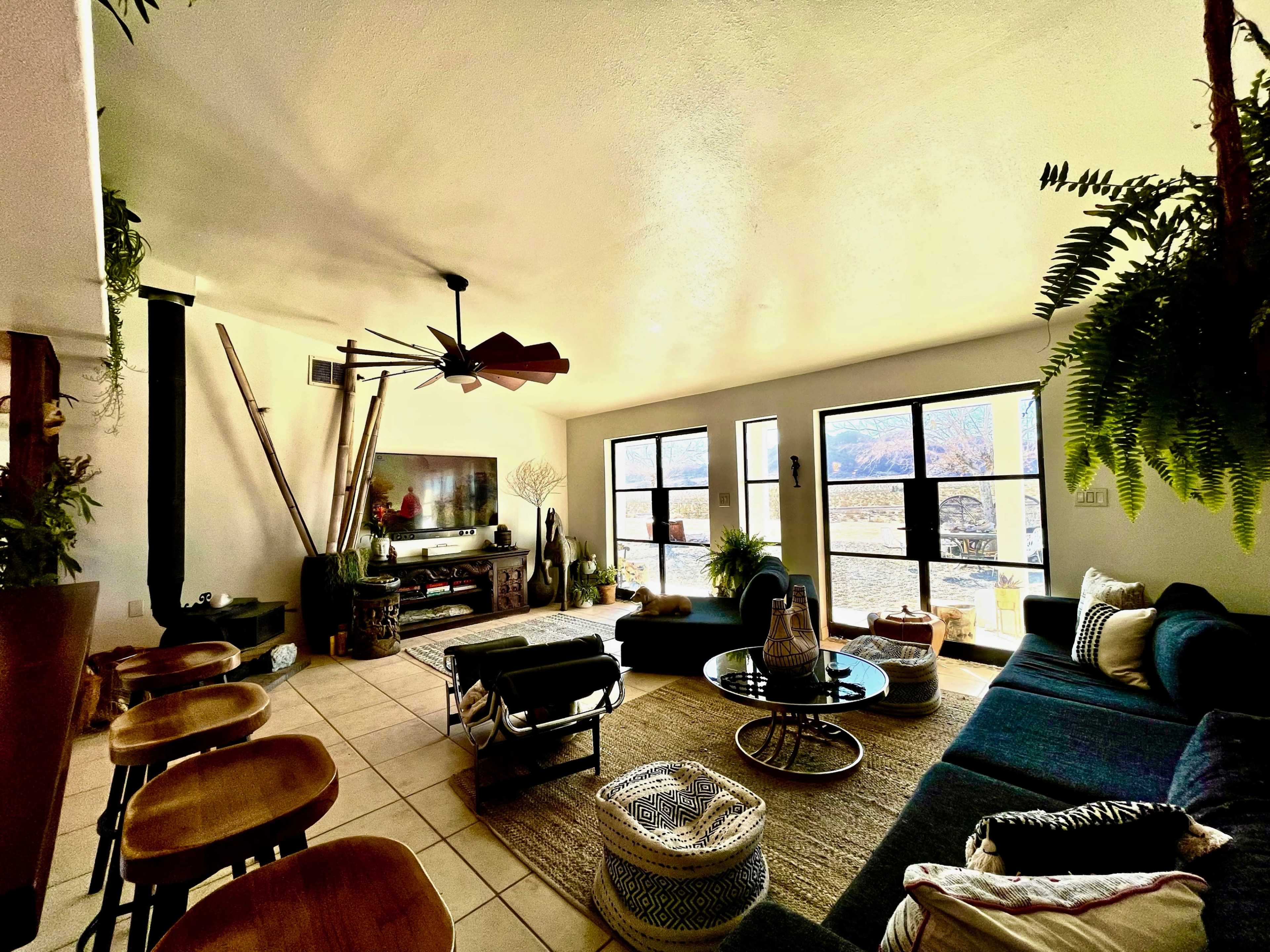 The image shows a bright living room furnished with a dark couch, a coffee table, a television, and large windows offering a view of the outdoors.