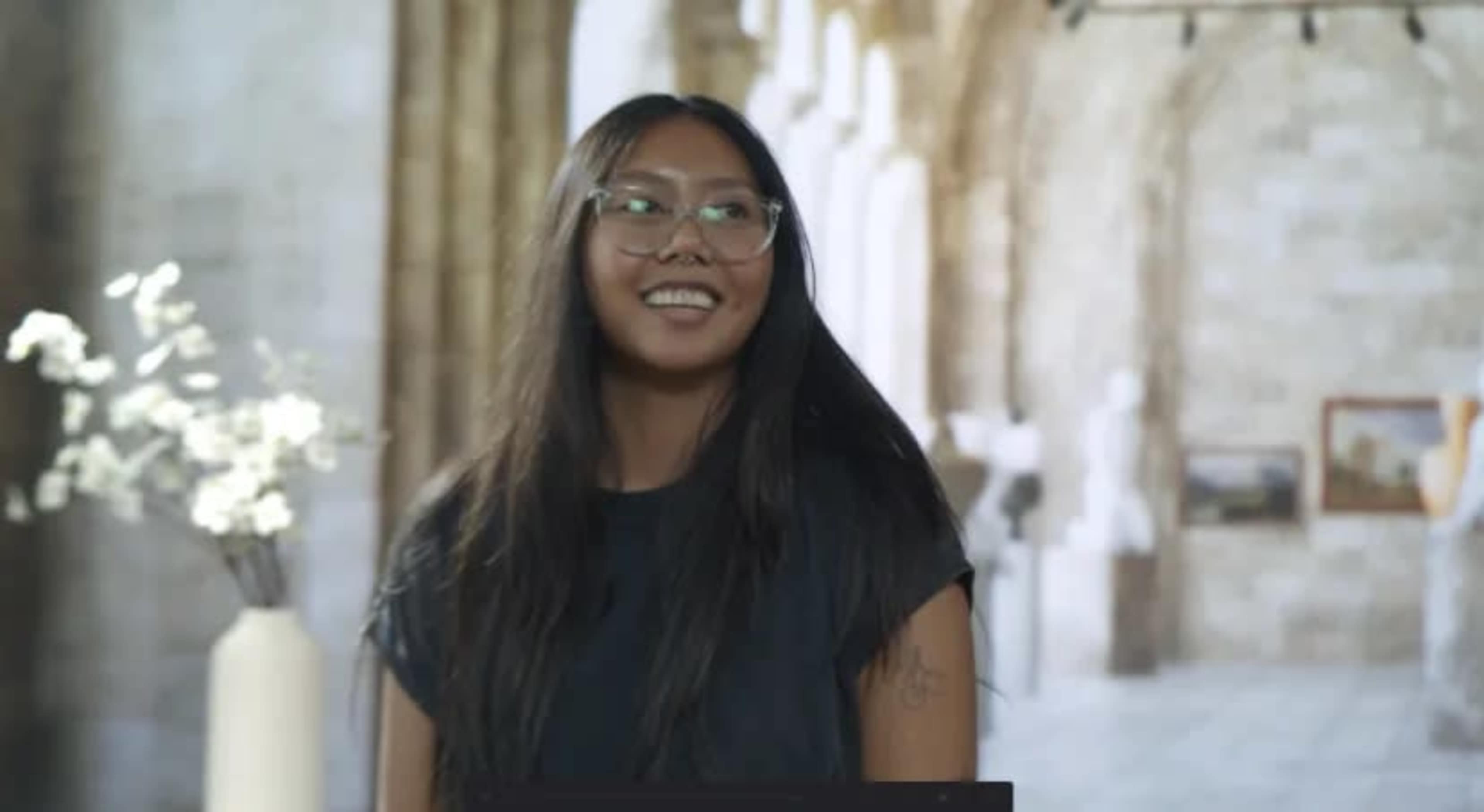 A woman with long black hair and glasses smiles while standing in a gallery space that features sculptures and paintings.