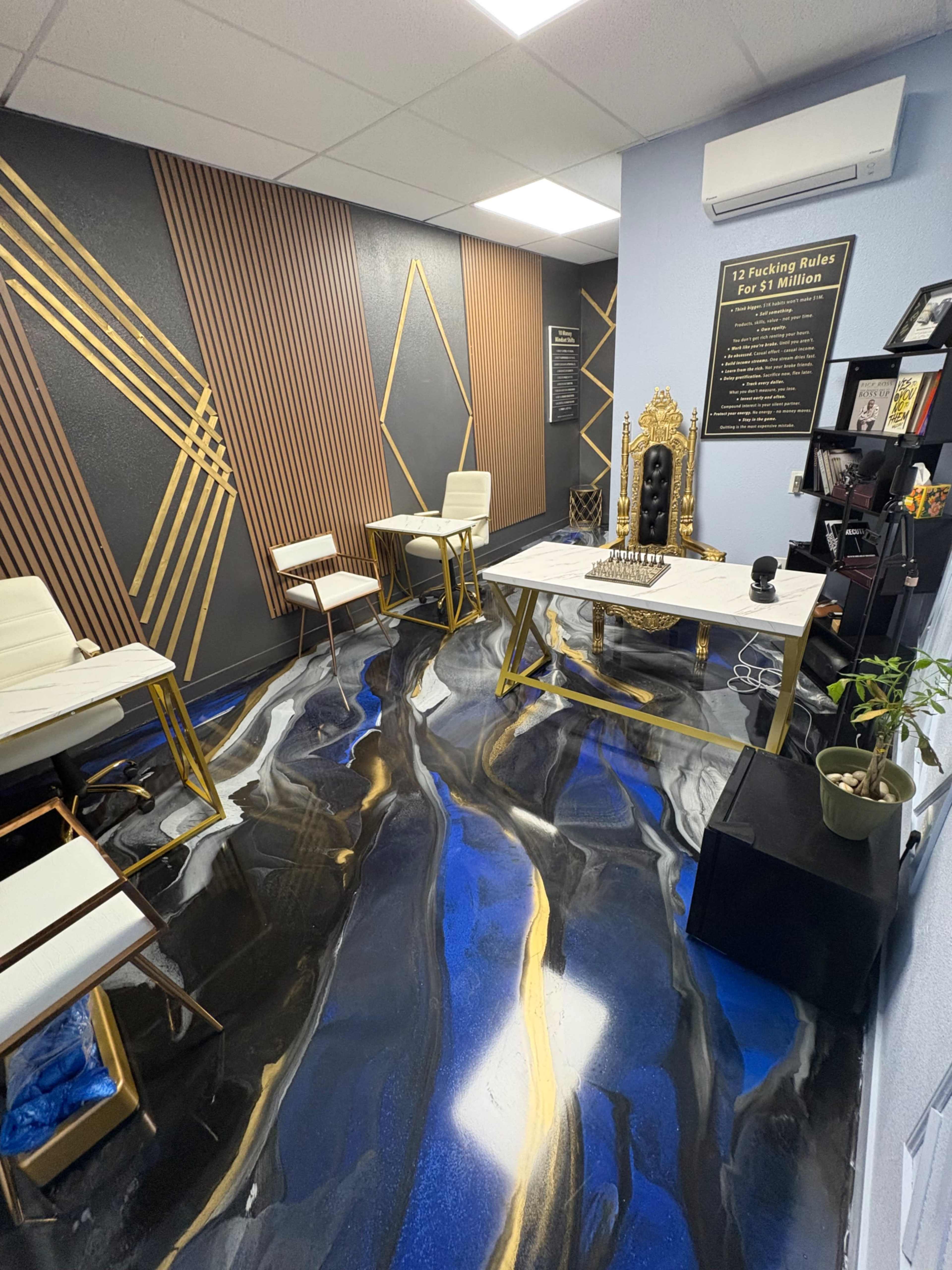 The image shows a modern office space with a blue and black epoxy floor, gold accents on the walls, and chic furniture, including two white chairs and a table.