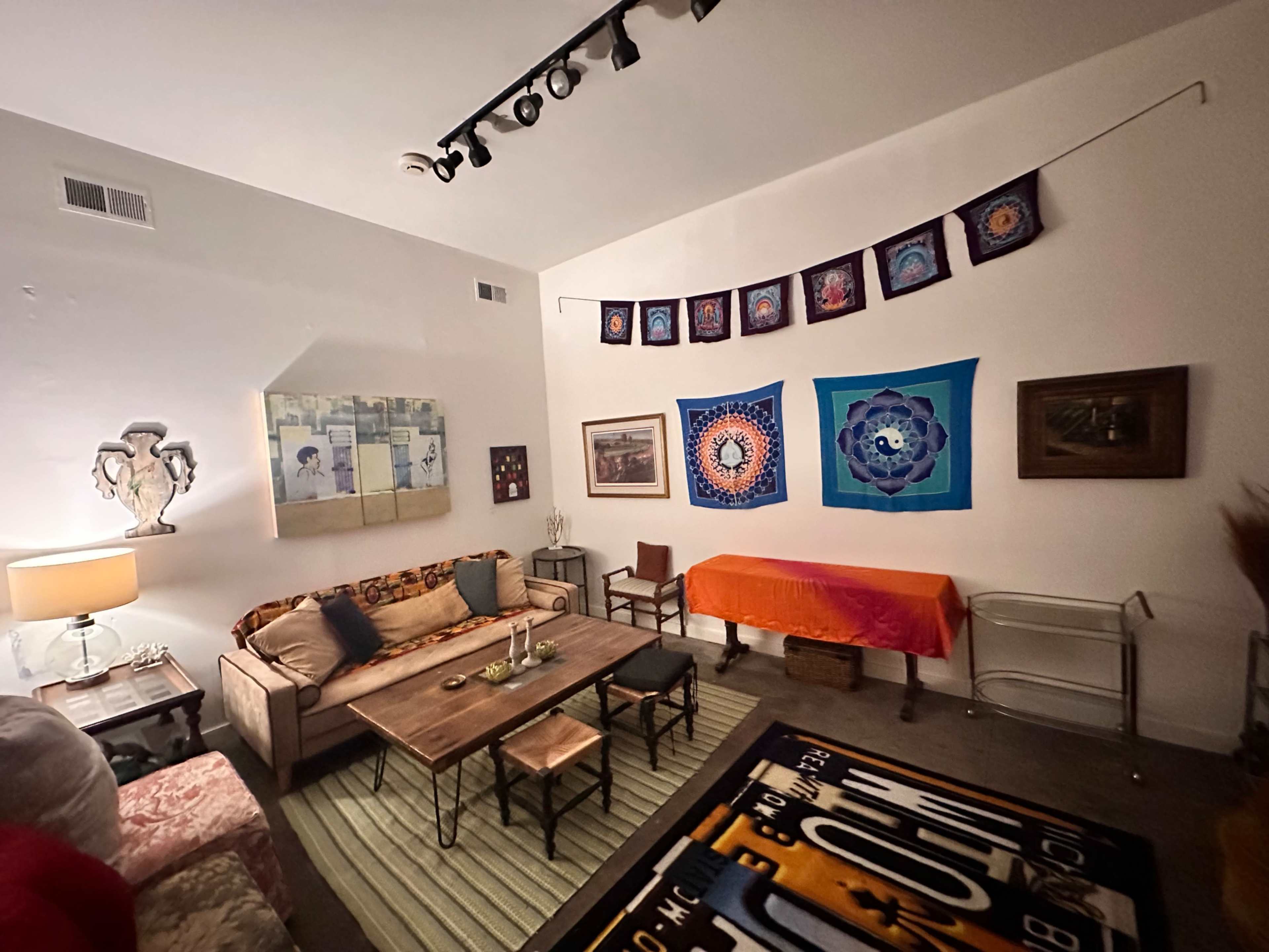 The image shows a cozy living room with a mix of colorful tapestries on the walls, a wooden coffee table centered among seating, and an orange tablecloth draped over a side table.