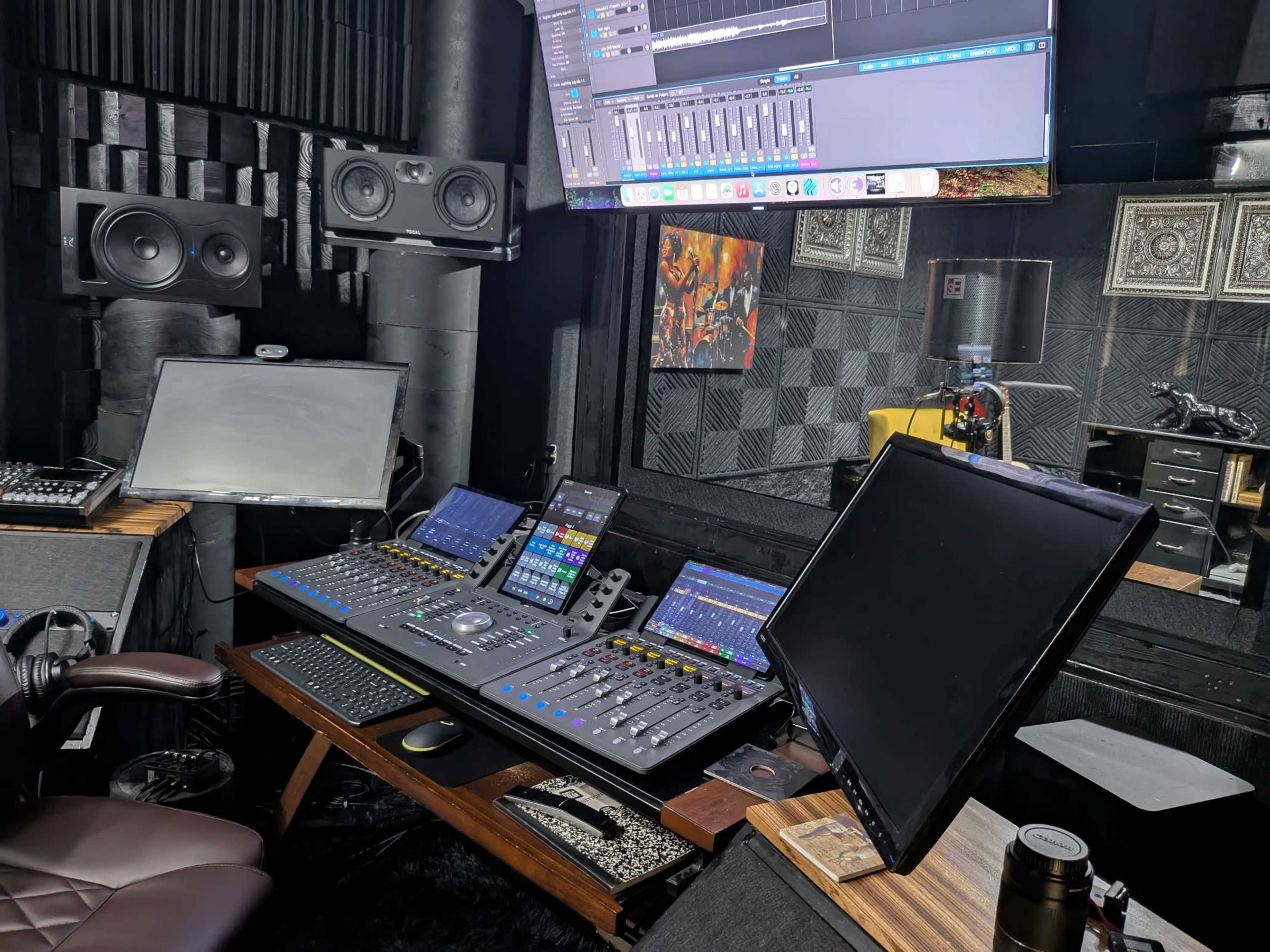 The image depicts a modern audio mixing studio with a digital mixing console, multiple monitors, and studio speakers arranged in a dark, acoustically treated room.