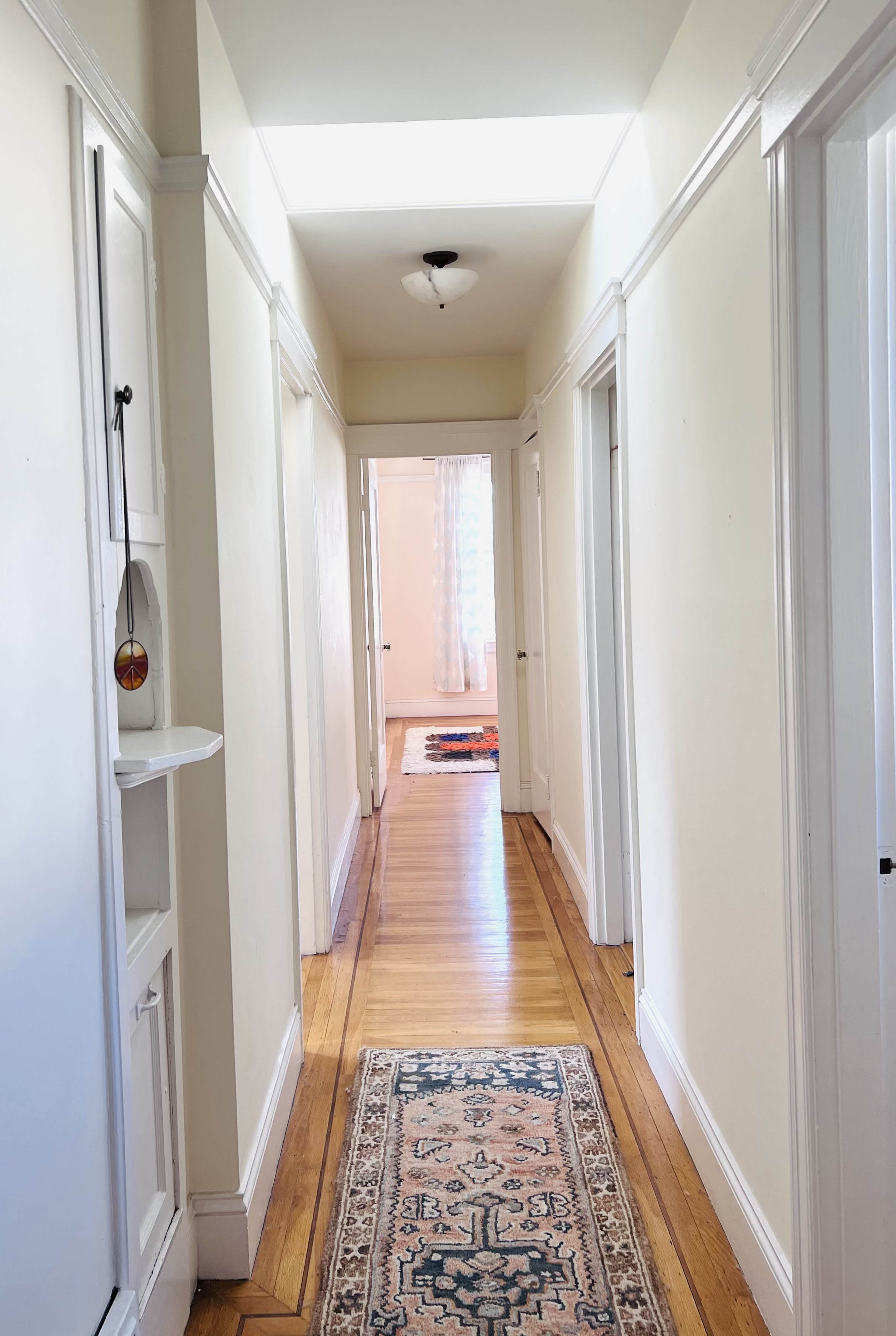 A long hallway with wooden flooring and a patterned rug leads to a room illuminated by natural light at the far end.