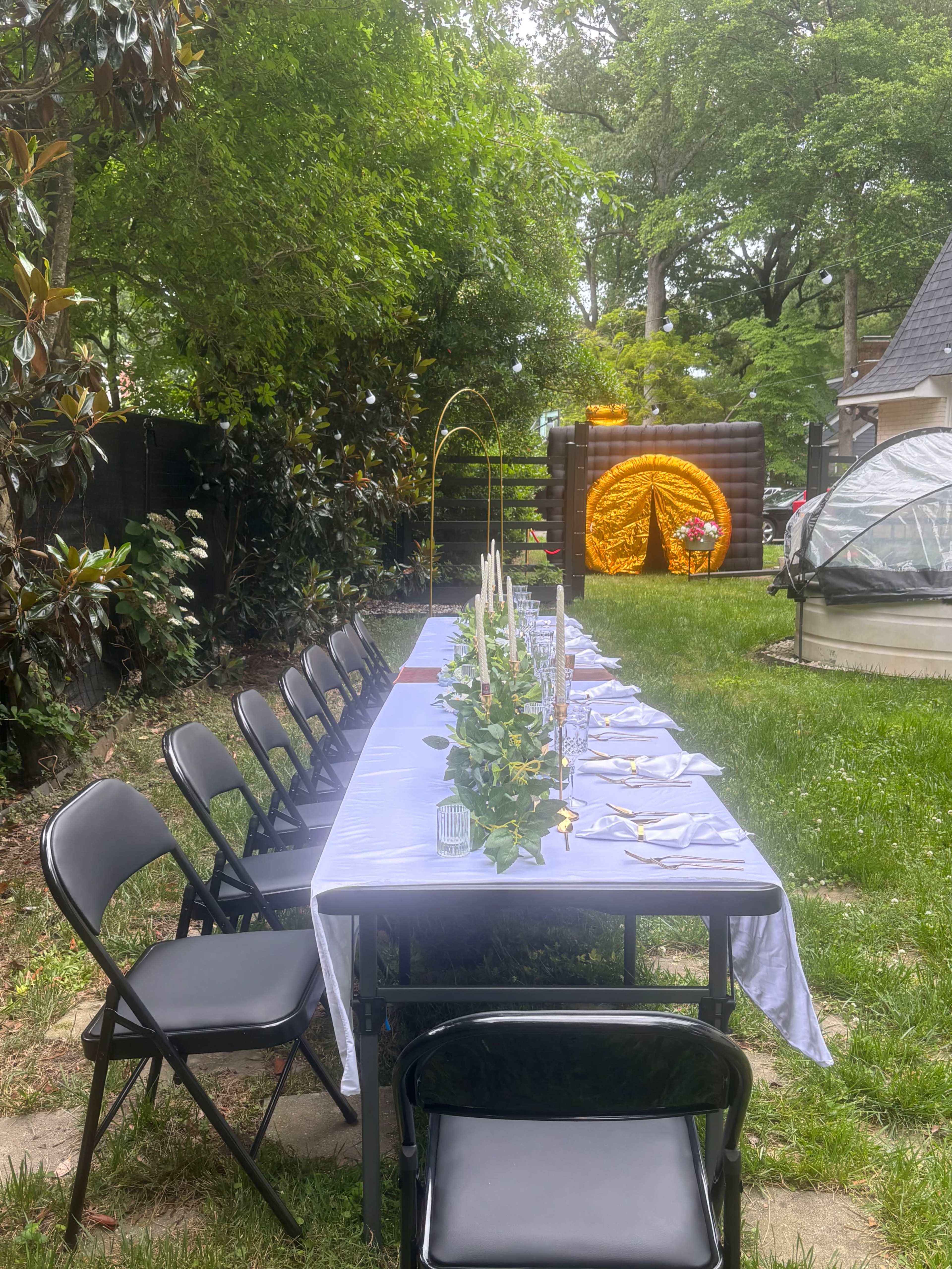 A long table is set up in a backyard, adorned with candles and greenery, with chairs arranged alongside it, and a decorative backdrop featuring a circular floral display nearby.