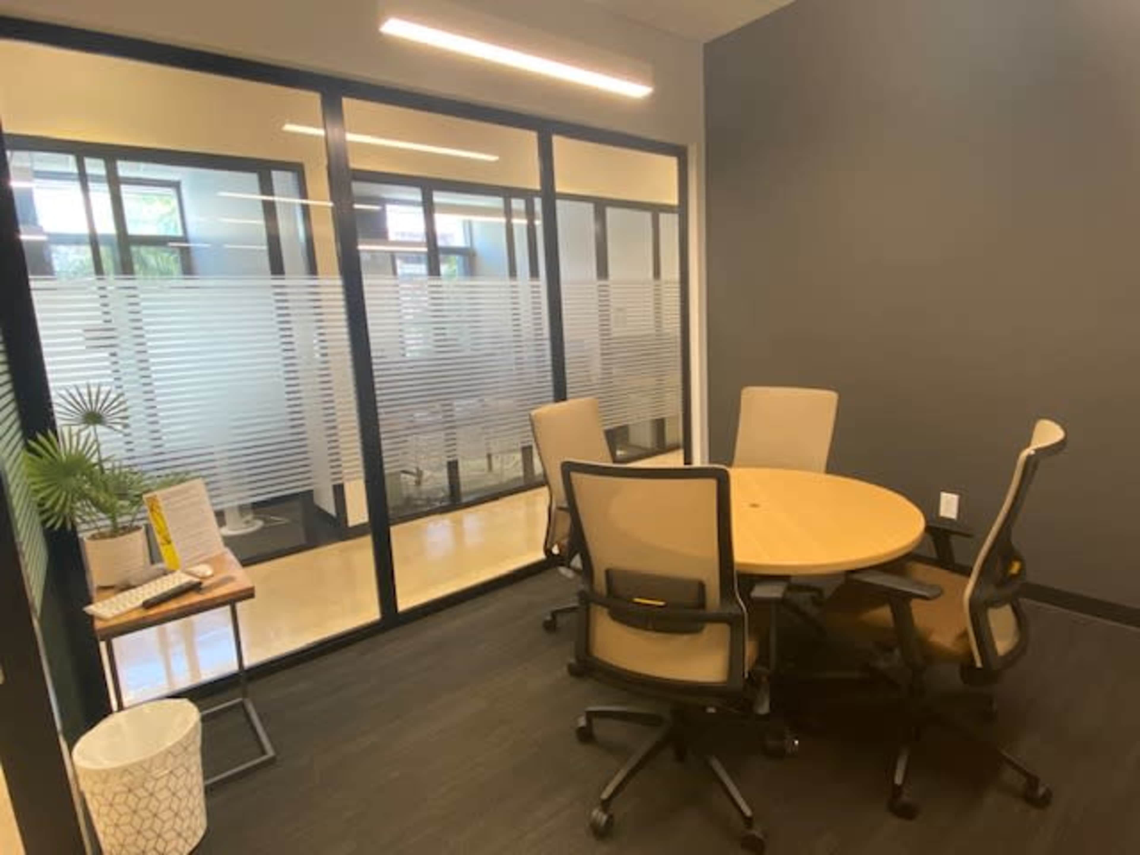 The image shows a small conference room with a round table and four chairs, adjacent to glass-paneled walls featuring frosted designs.