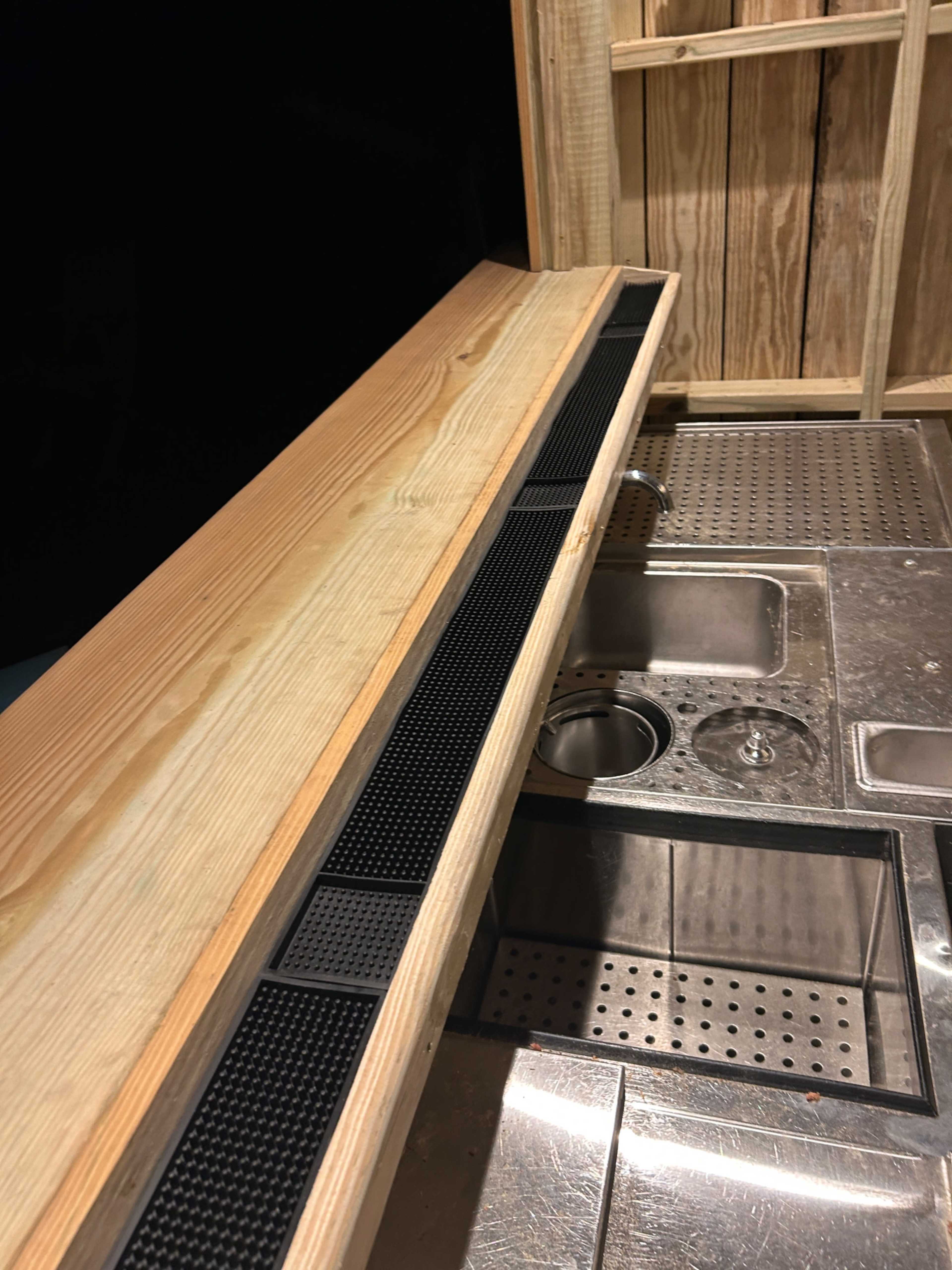 The image shows a wooden counter with a built-in drainage system and a stainless steel bar setup beneath it.