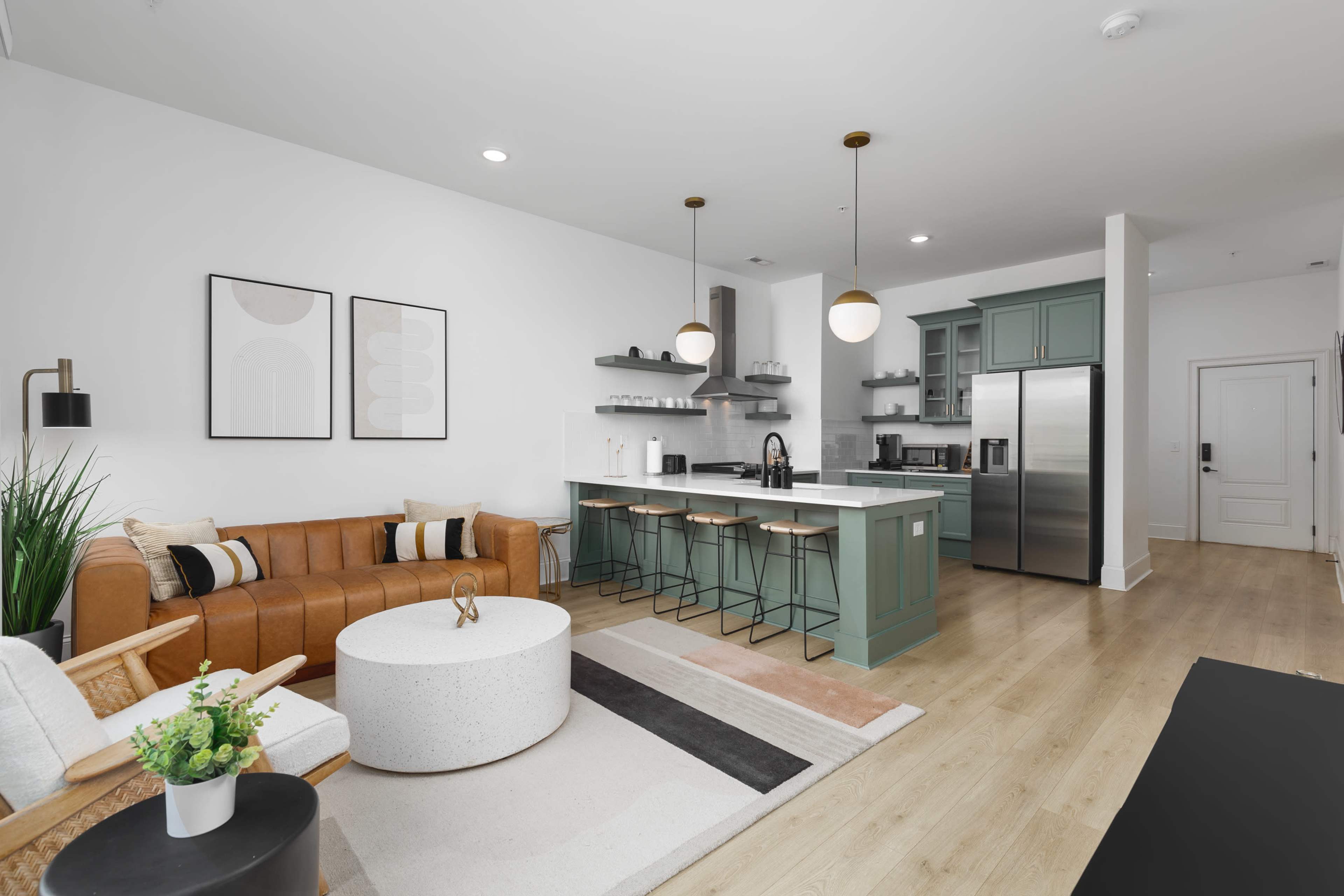 A modern living space features a brown leather sofa, a round white coffee table, and an open kitchen with green cabinetry and stainless steel appliances.