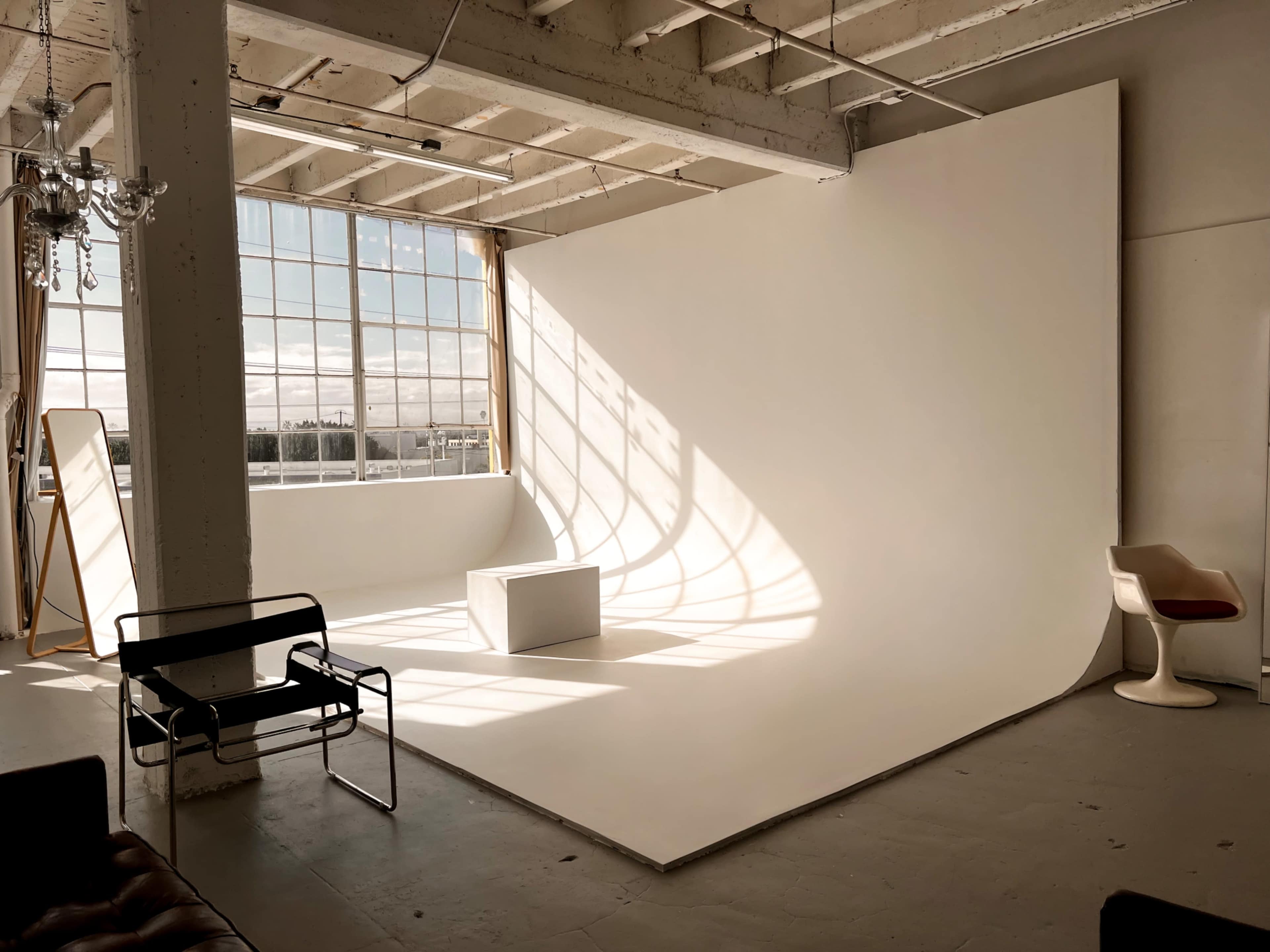 The image shows a spacious photography studio with a large white backdrop, natural light streaming through tall windows, and minimal furniture including a chair and a black metal chair.