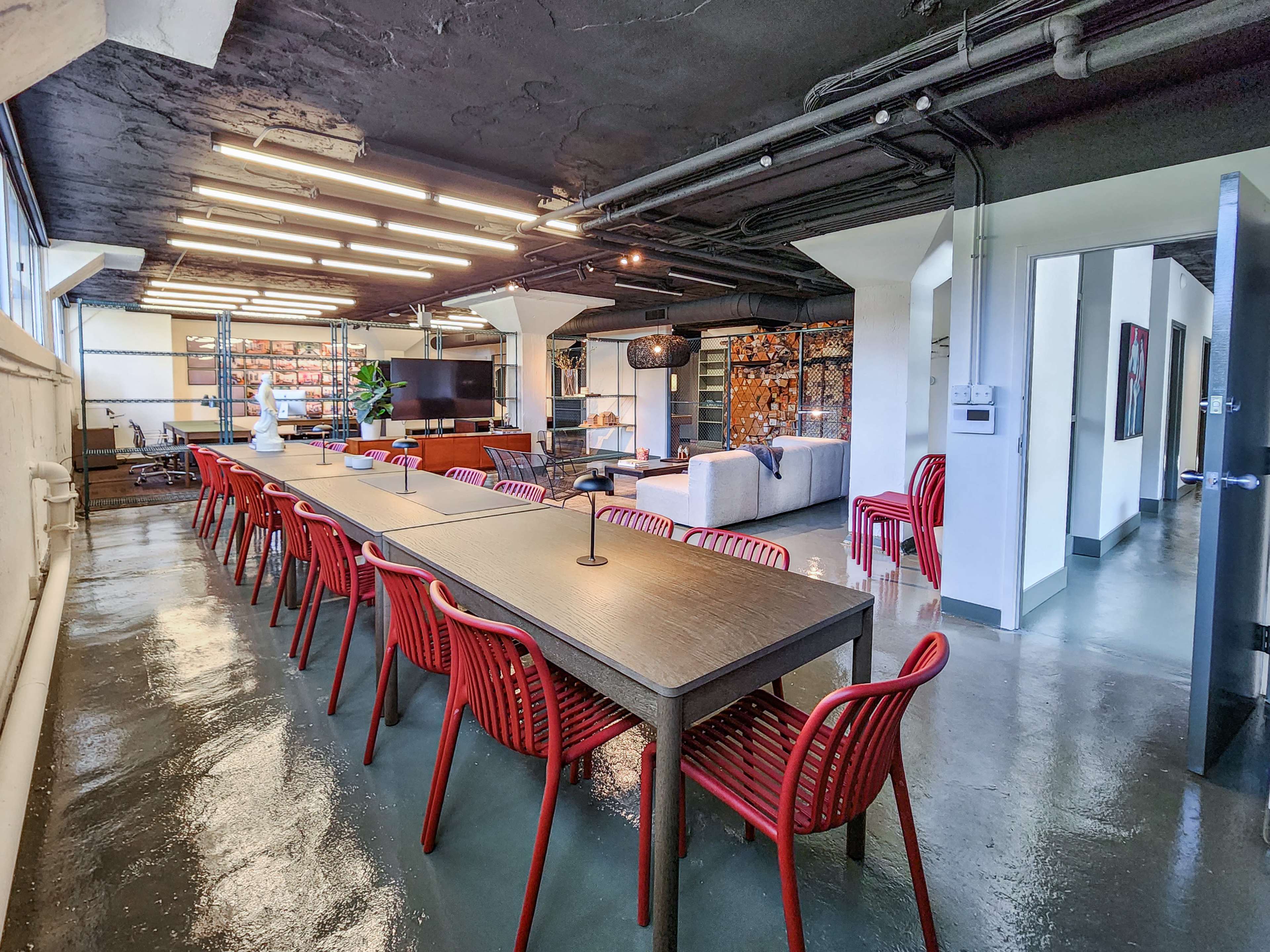 A long dining table with red chairs occupies a modern, open workspace featuring a lounge area and exposed ceiling pipes.