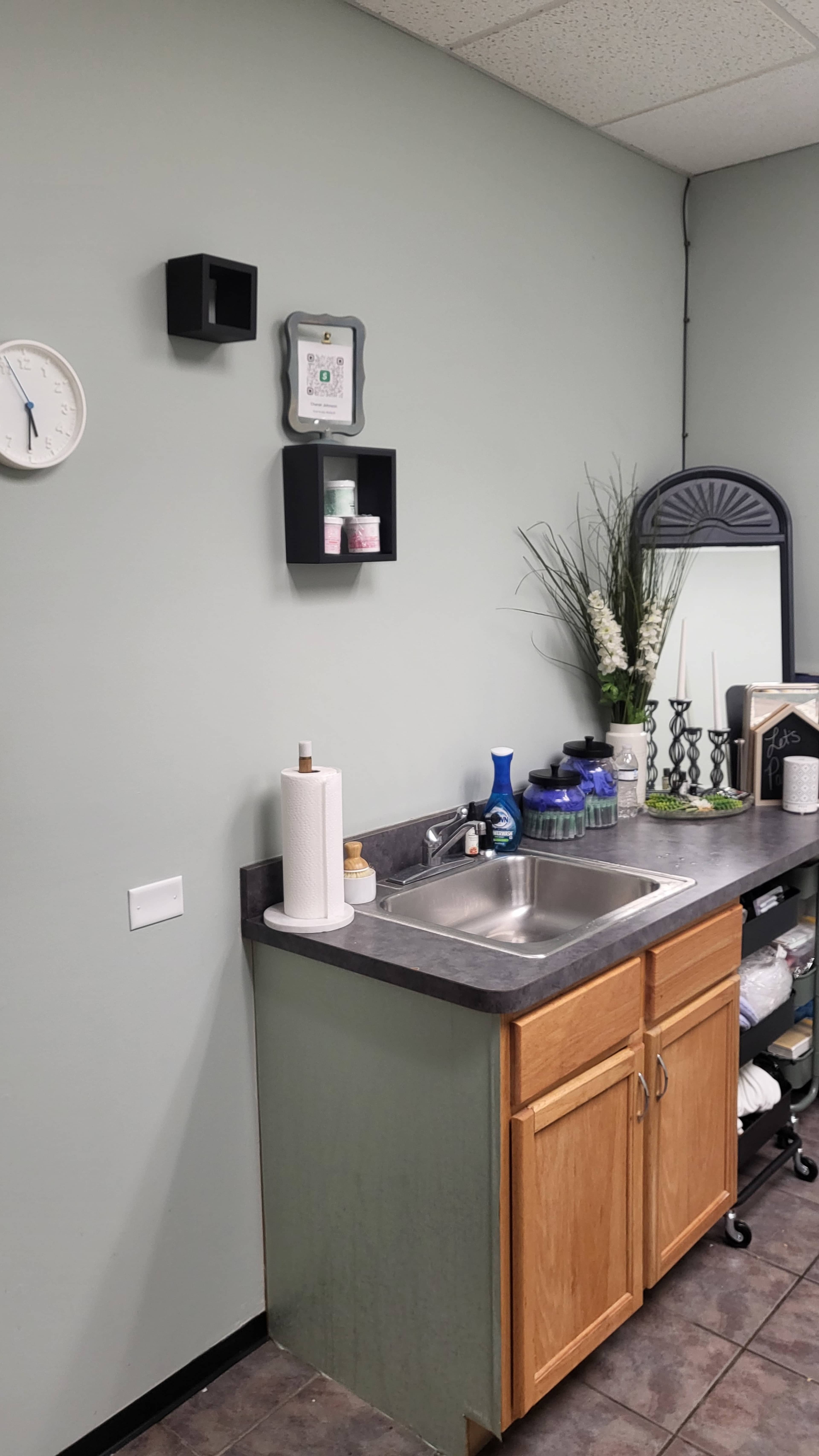 The image shows a small room with a sink and wooden cabinets beneath a gray countertop, accompanied by wall shelves and a mirror on the right.