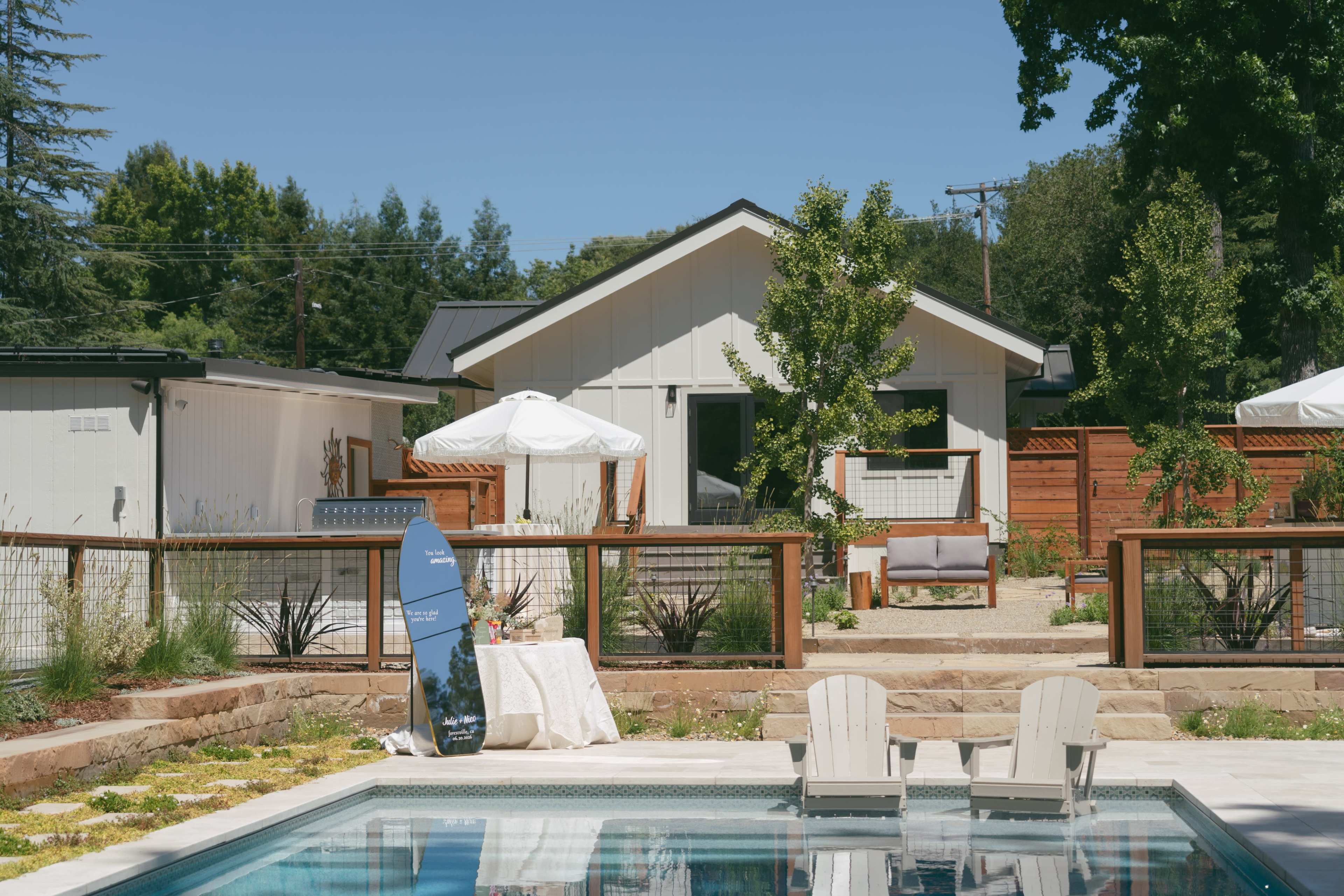 A modern backyard features a swimming pool, a wooden deck, a shaded seating area with an umbrella, and a house in the background surrounded by greenery.