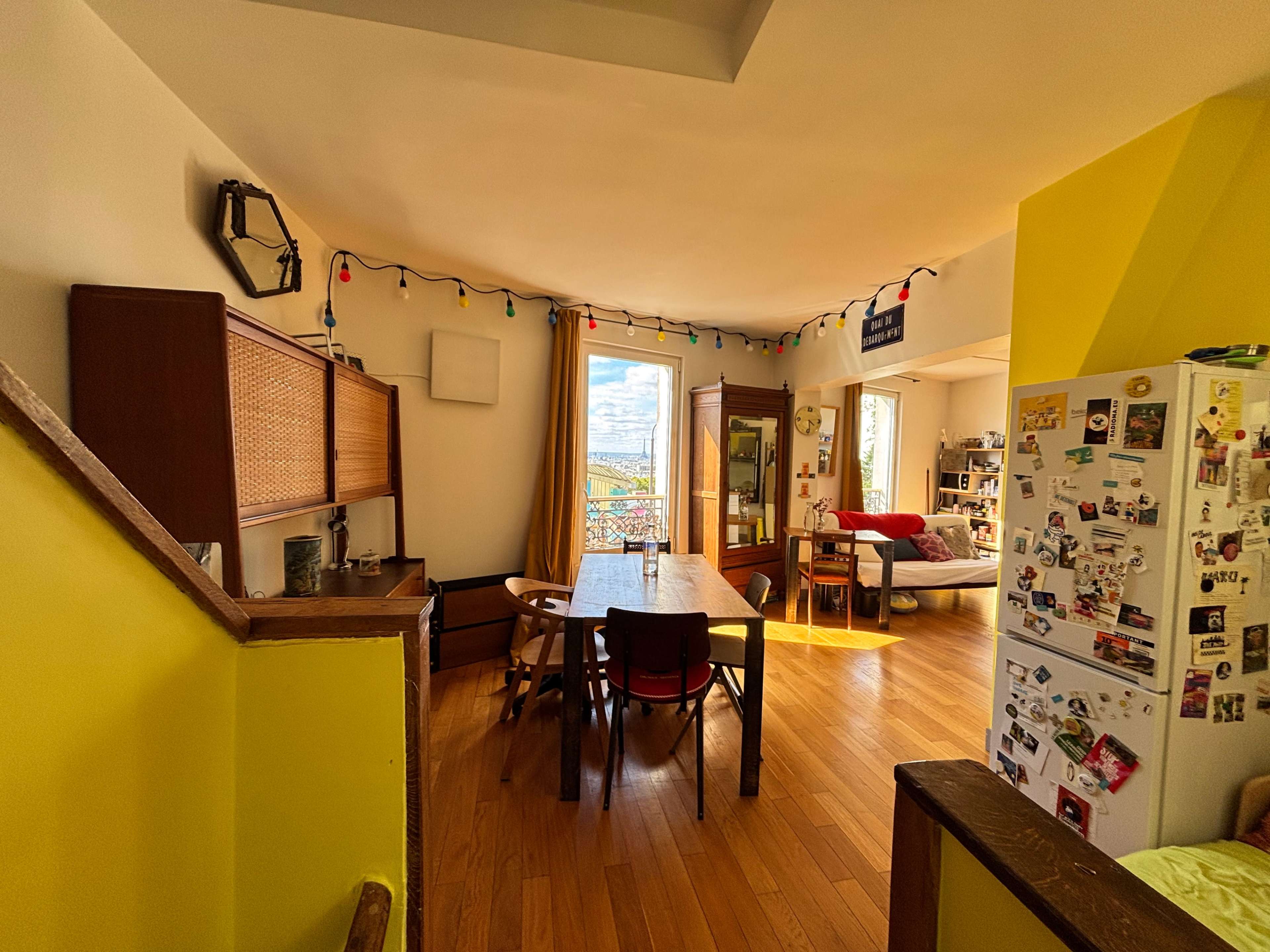 A bright room features a dining table, chairs, a refrigerator covered in magnets, and a window with a view of the outdoors.