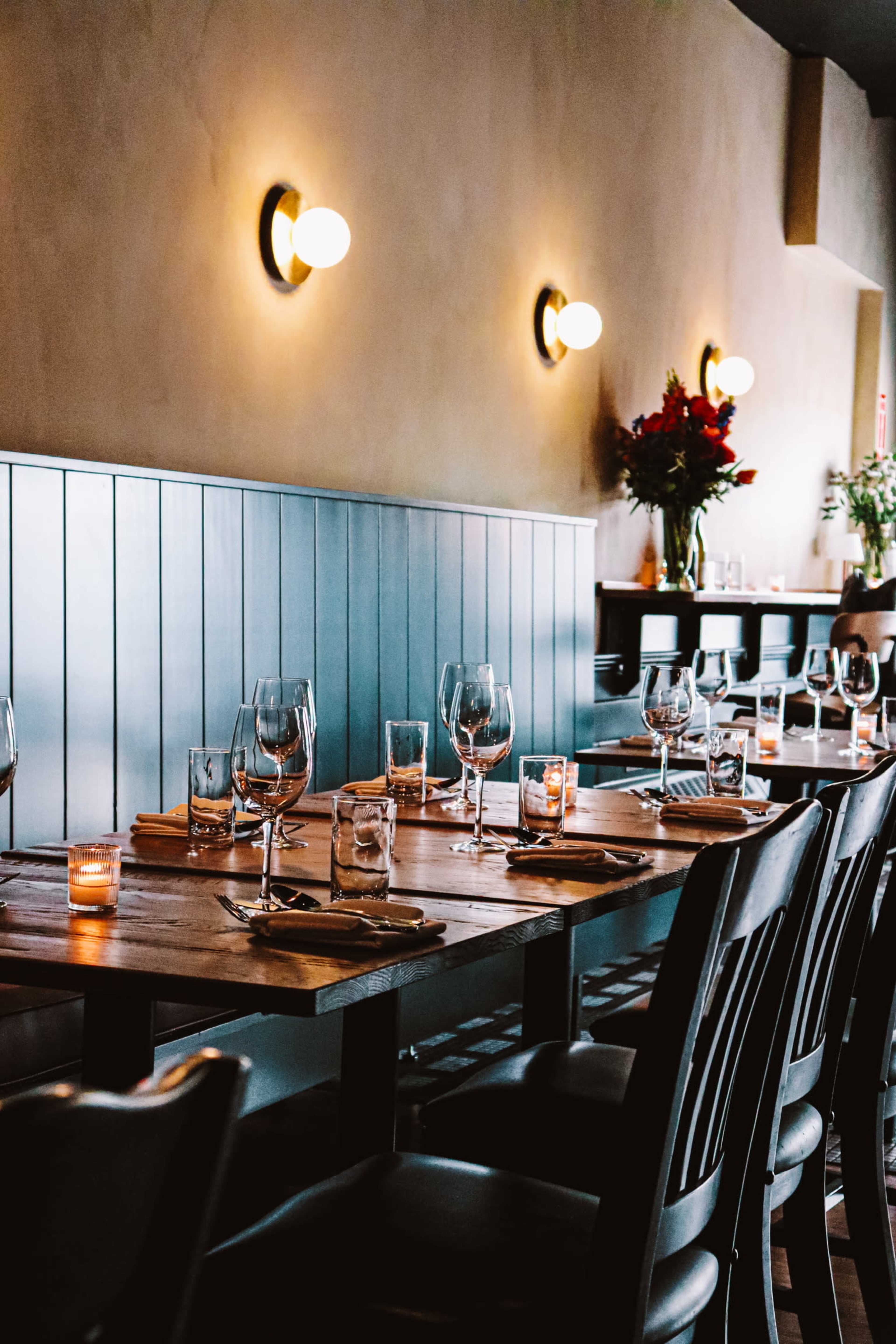 The image shows a dimly lit restaurant interior with wooden tables set for dining, featuring wine glasses, water glasses, and candles, along a blue-paneled wall.