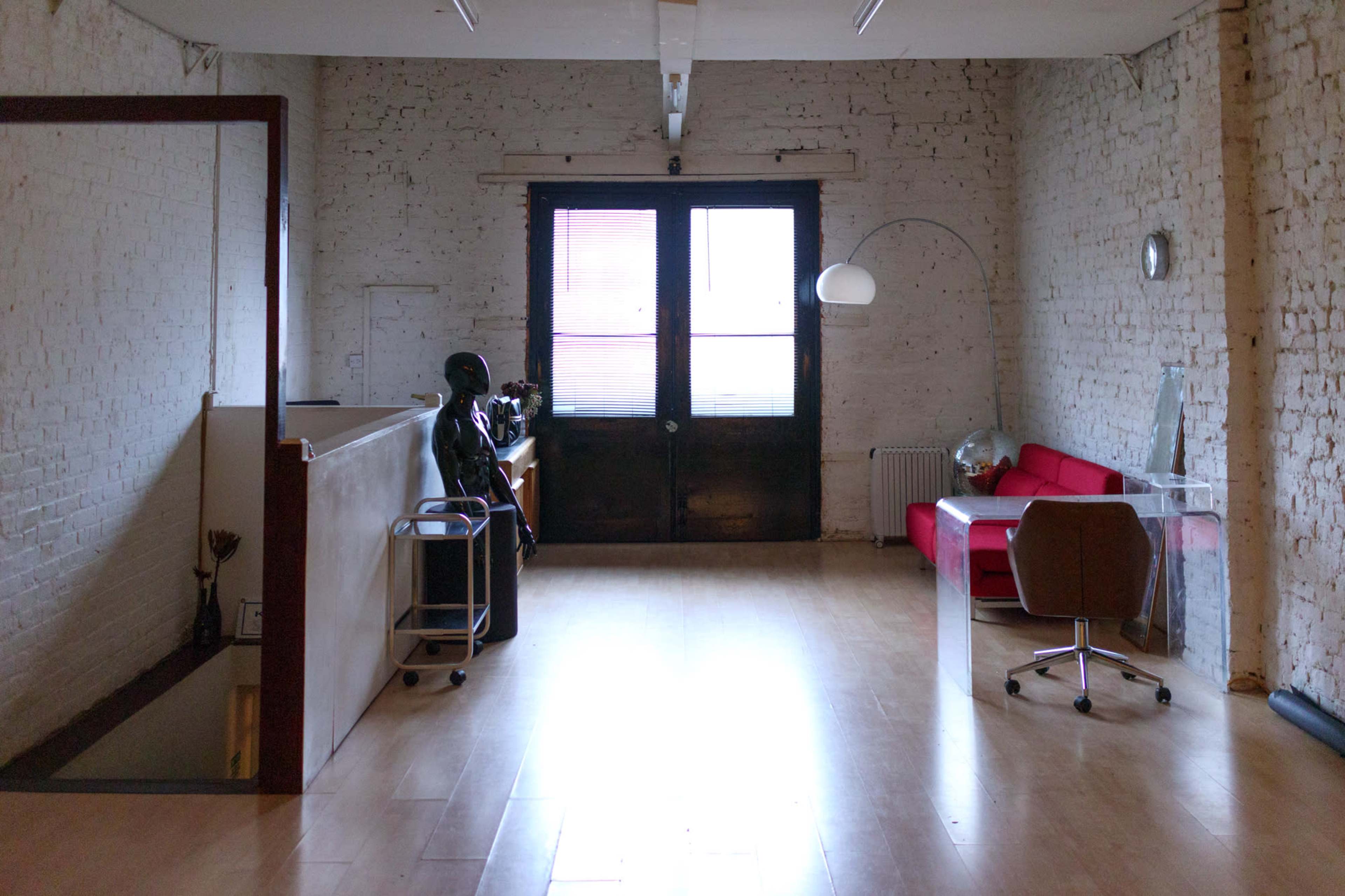 A minimalist interior space with a wooden floor, white brick walls, a red sofa, and a large window with a dark wooden door.